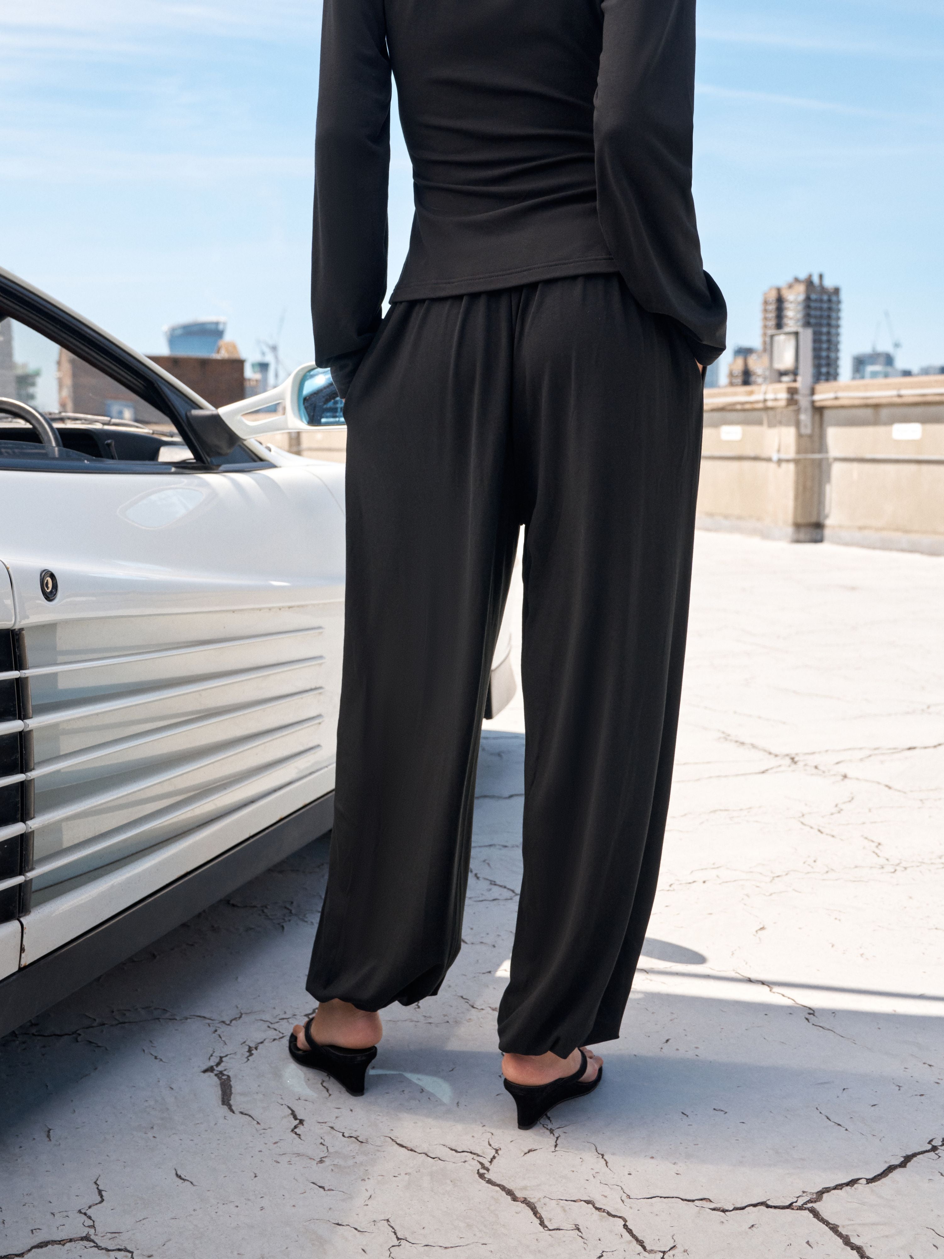 A person wearing a black long-sleeve top and loose black wide-leg pants, standing outdoors beside a white car on a cracked concrete surface with a city skyline in the background.