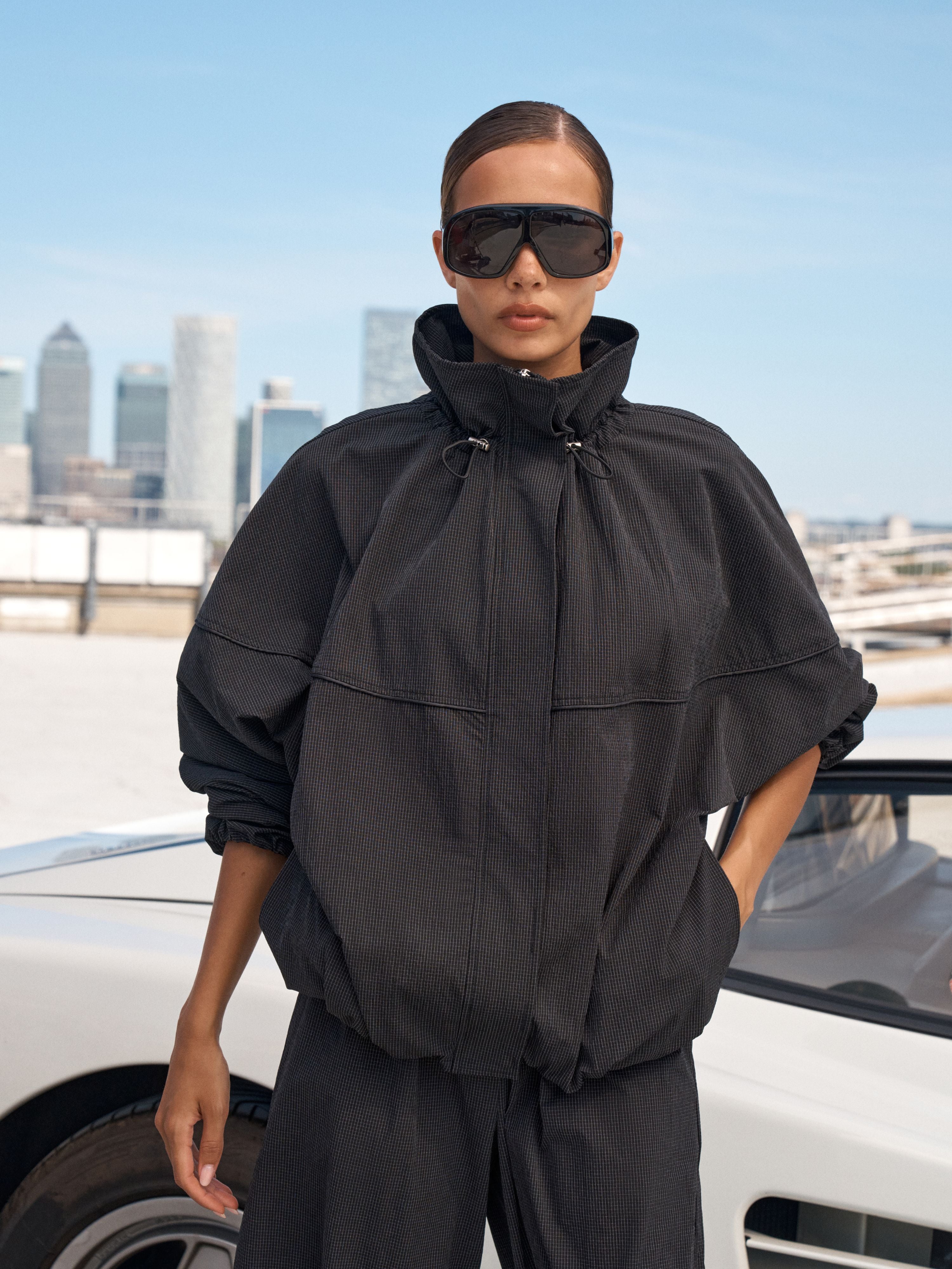 A model wearing a black oversized zip-up jacket with a high collar and matching black pants, standing outdoors next to a white car with a city skyline in the background.