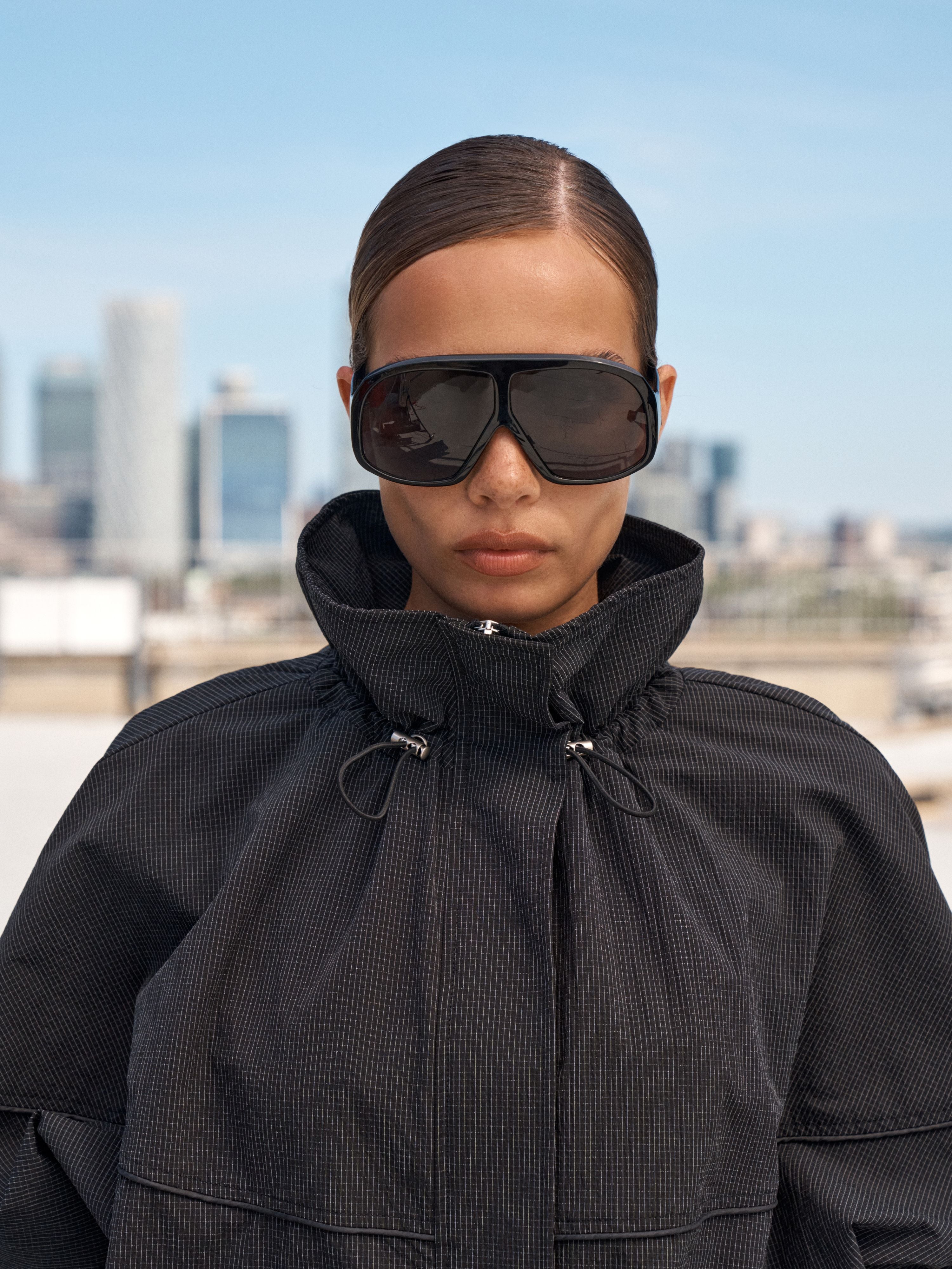 A model wearing oversized black shield sunglasses and a black textured high-collar jacket, photographed outdoors with a blurred city skyline and blue sky in the background.