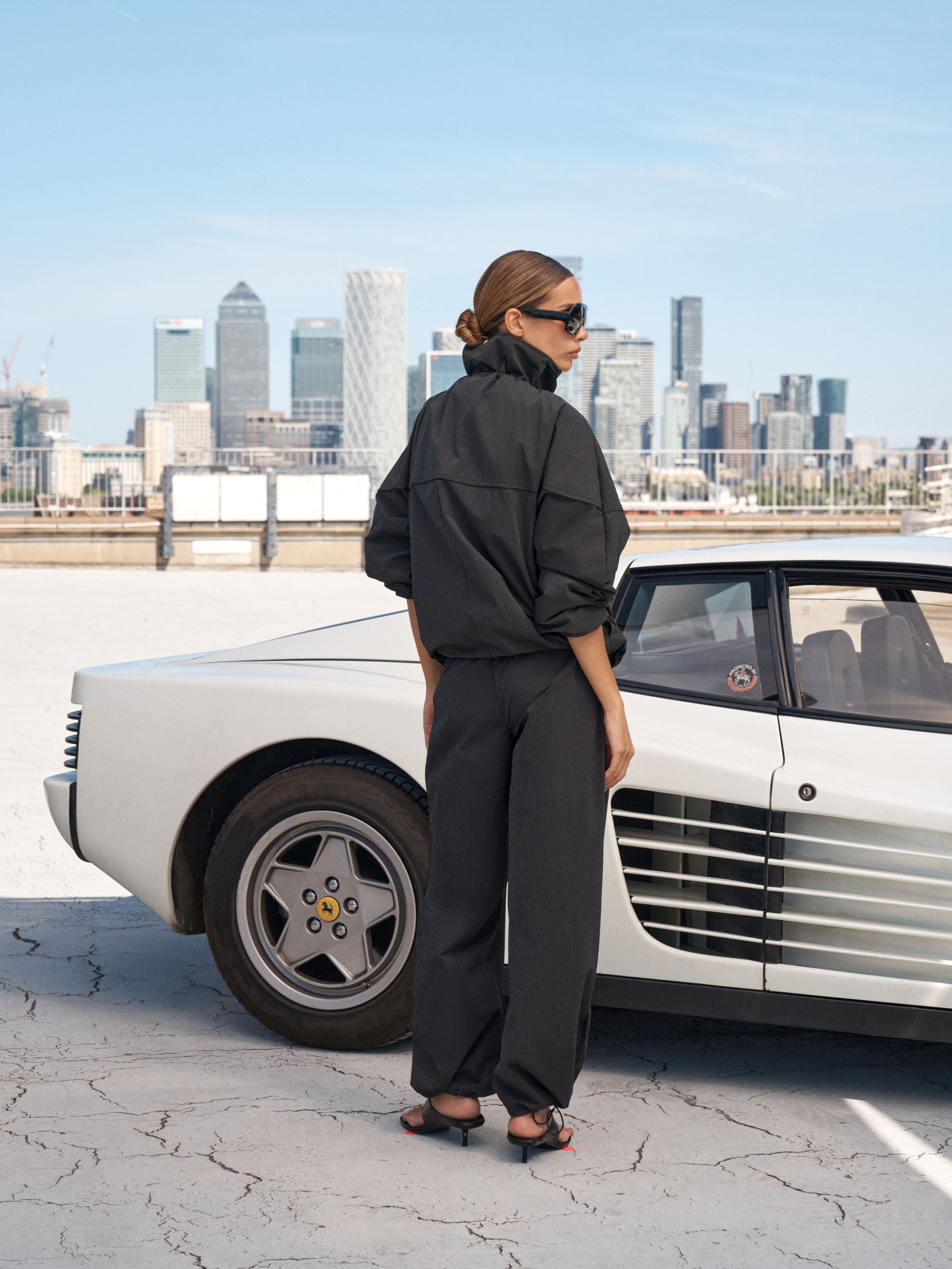 A woman wearing a black oversized jacket and matching wide-leg trousers stands beside a white classic sports car, with a city skyline in the background under a clear blue sky.