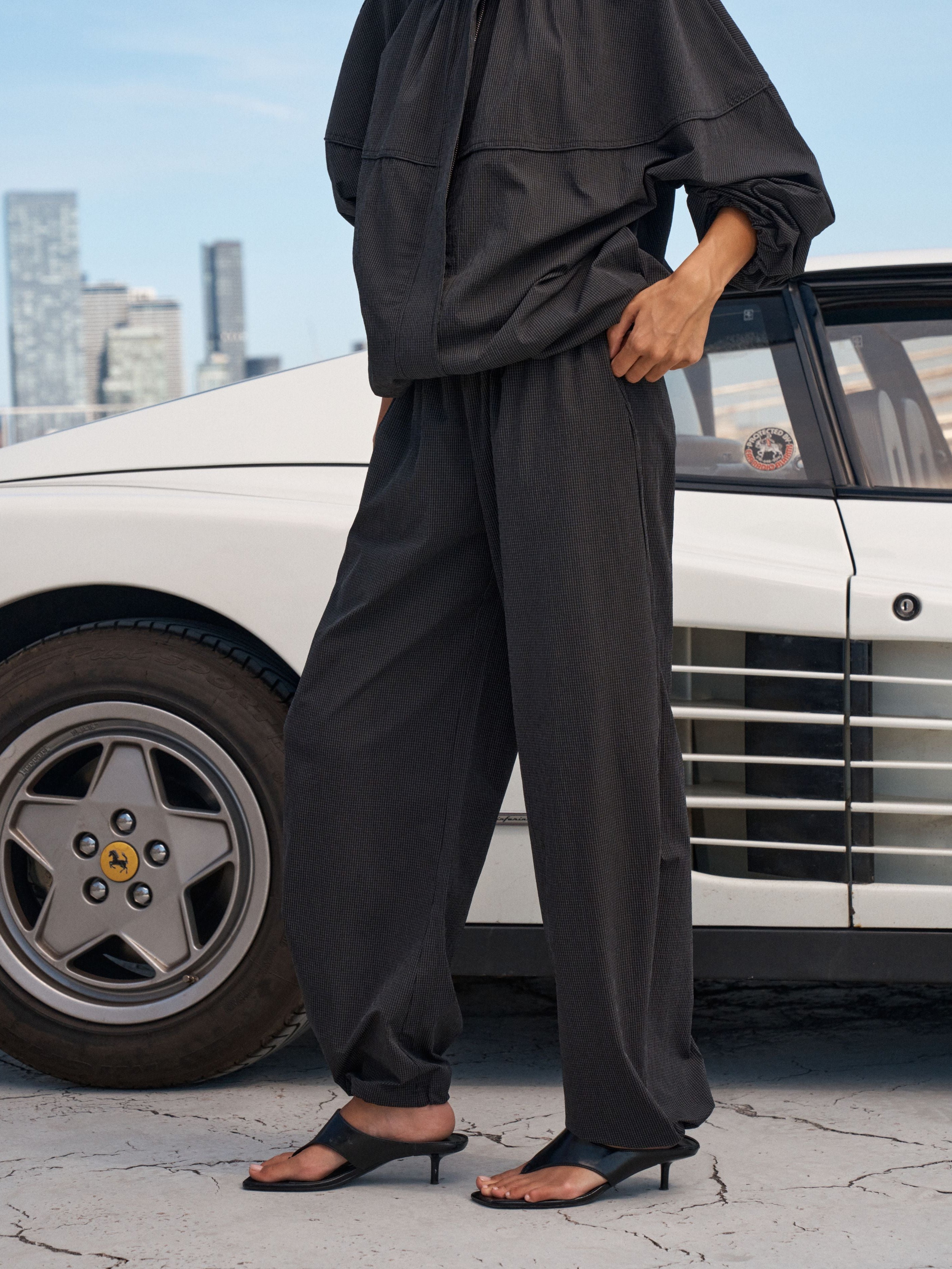 A person wearing loose black draped pants and a matching black top stands beside a white classic car on a cracked pavement with a city skyline in the background.