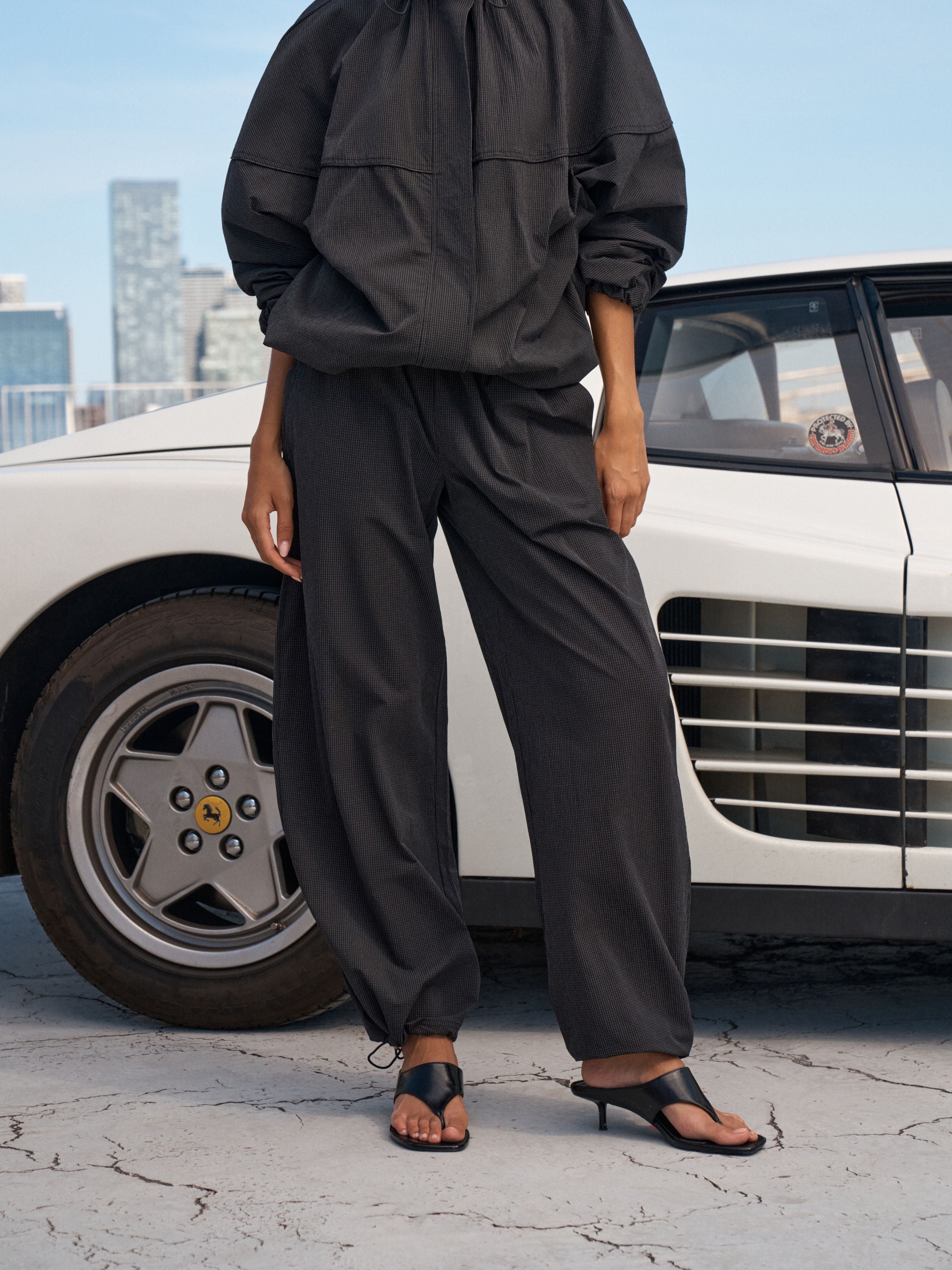 A model wearing a black lightweight two-piece set consisting of a cropped pullover top and matching wide-leg trousers, standing outdoors next to a white classic car.