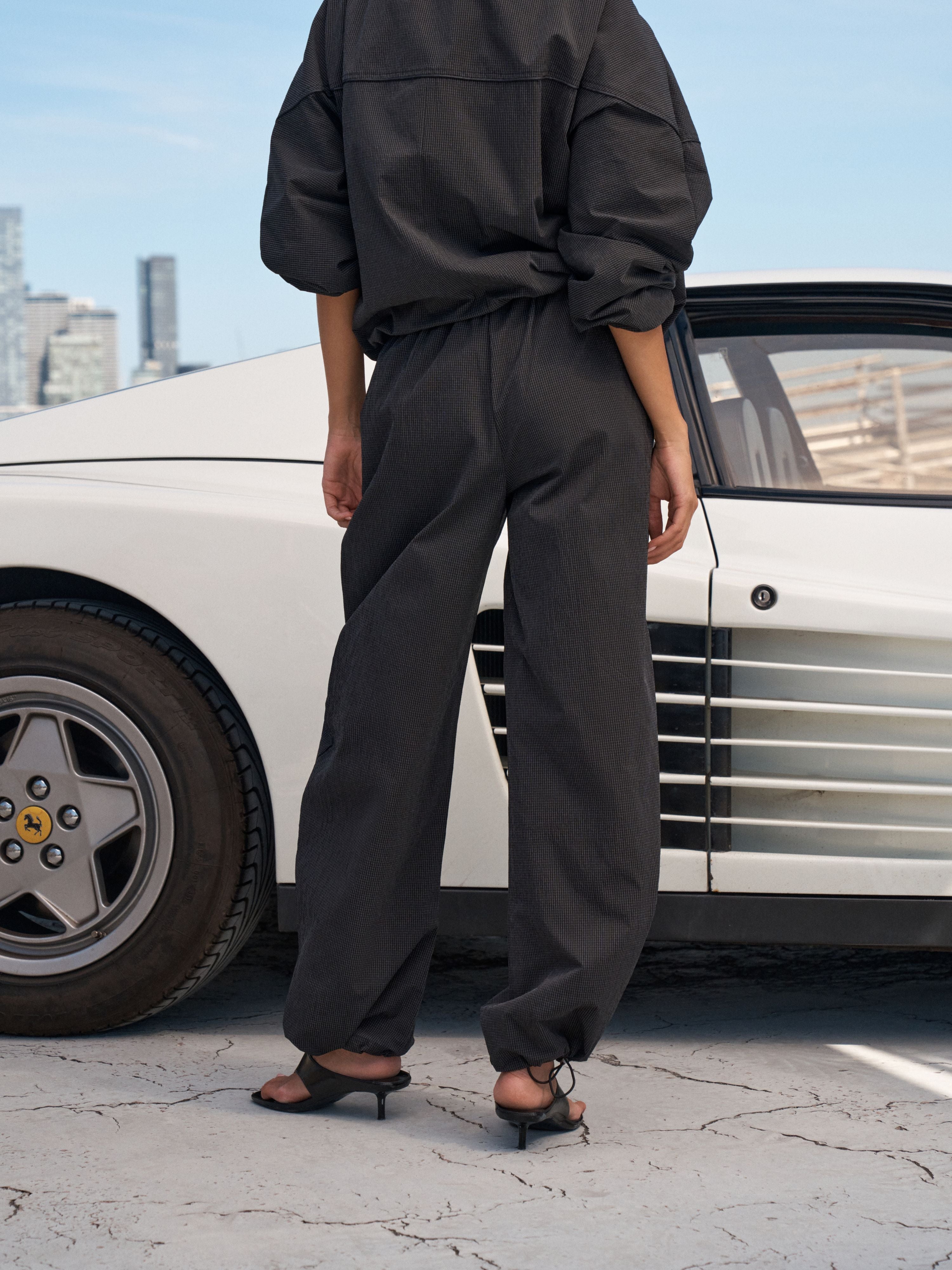 A model from behind wearing a black oversized long-sleeve top and matching wide-leg trousers, standing next to a white classic sports car on a city street.