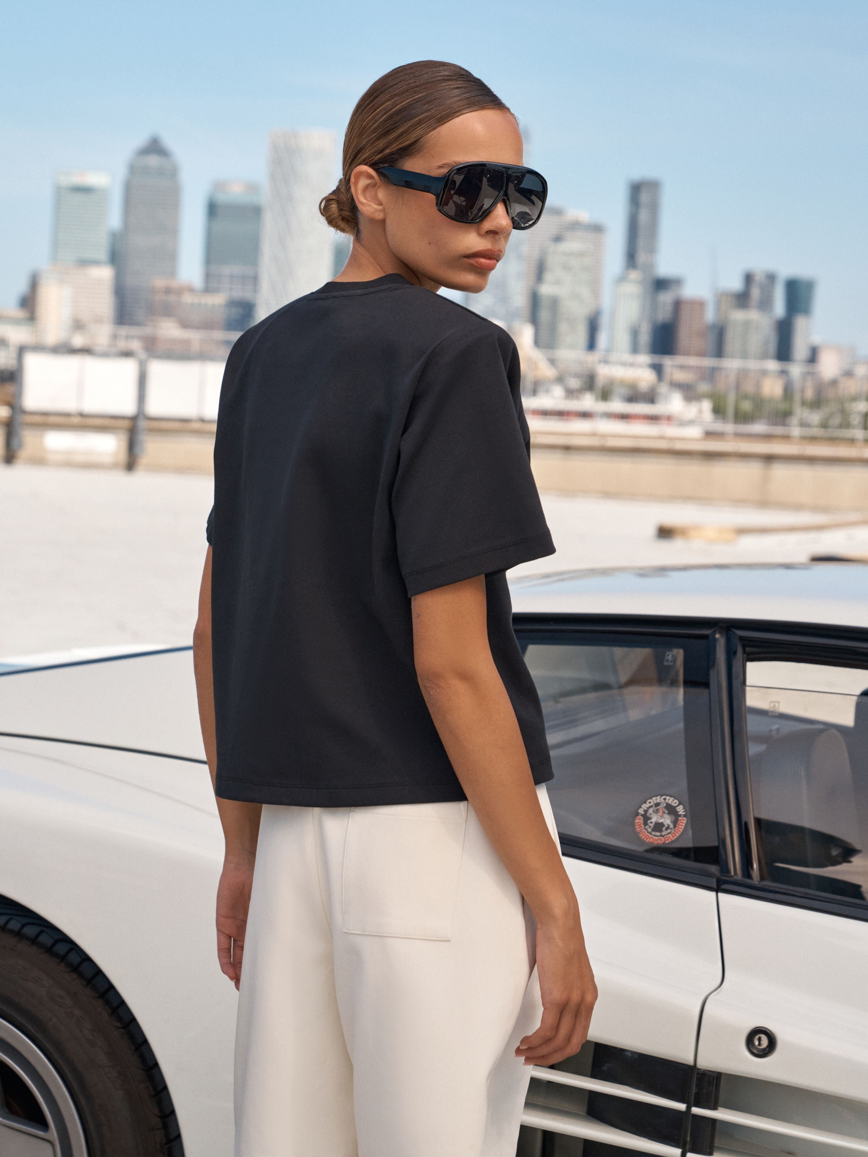 A woman wearing a black short-sleeve crop top and white trousers stands next to a white classic car with a city skyline in the background.
