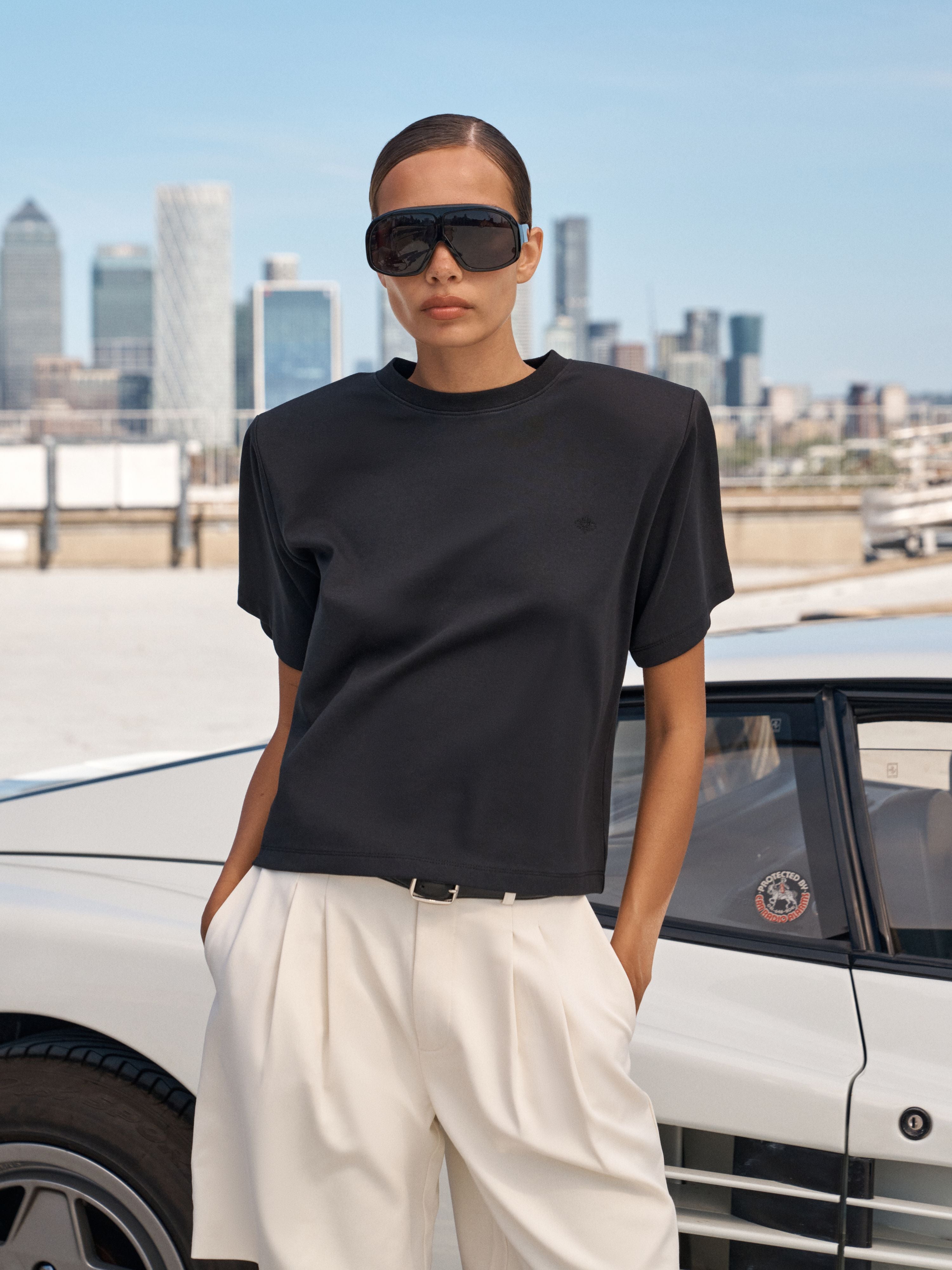 A model wearing a black short-sleeve crewneck t-shirt and white wide-leg trousers, standing outdoors next to a white car with a city skyline in the background.