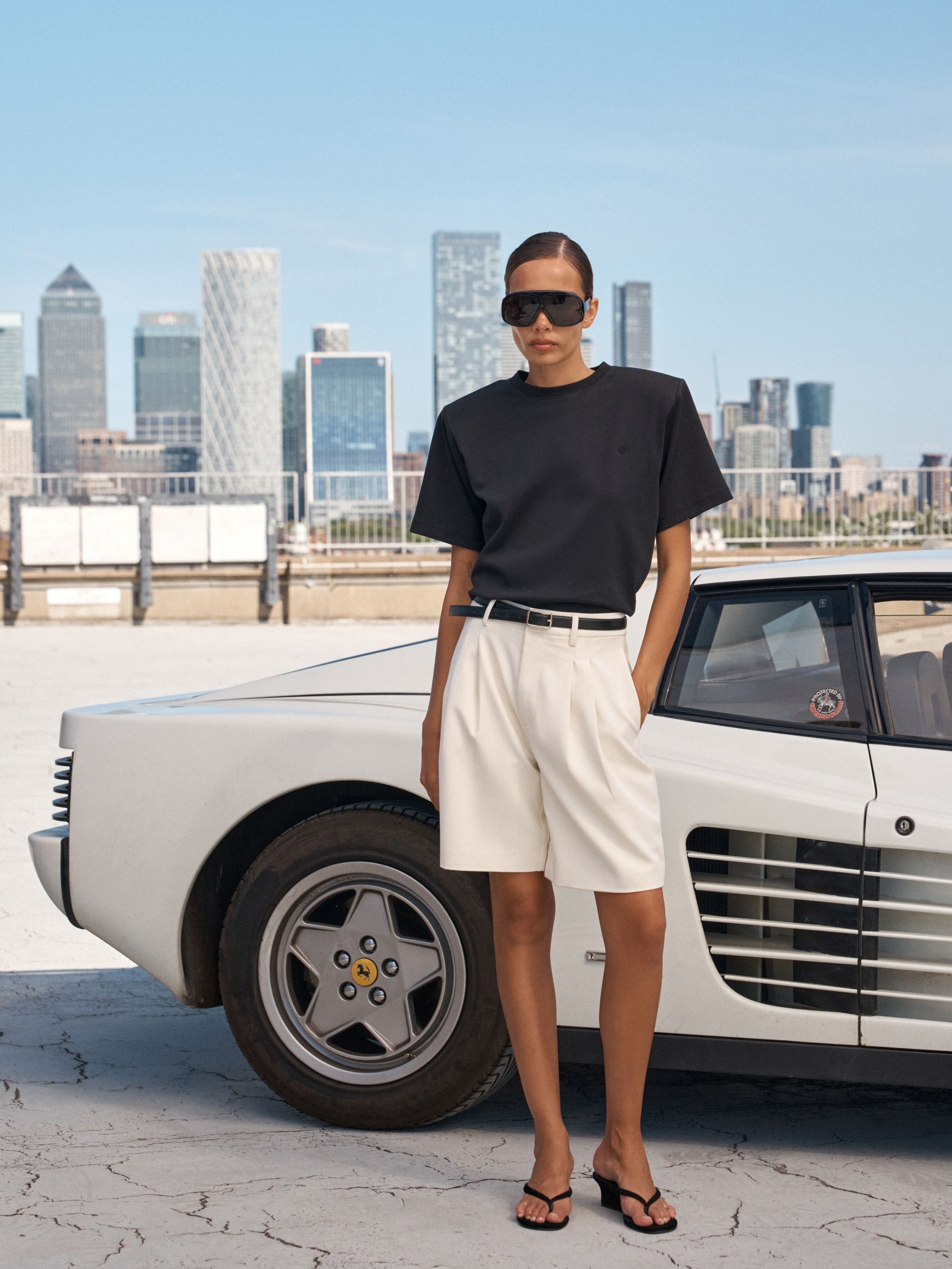 A model stands outdoors next to a white classic car, wearing a black short-sleeve t-shirt, off-white shorts, a thin black belt, and black sandals. The background features a city skyline under a clear blue sky.