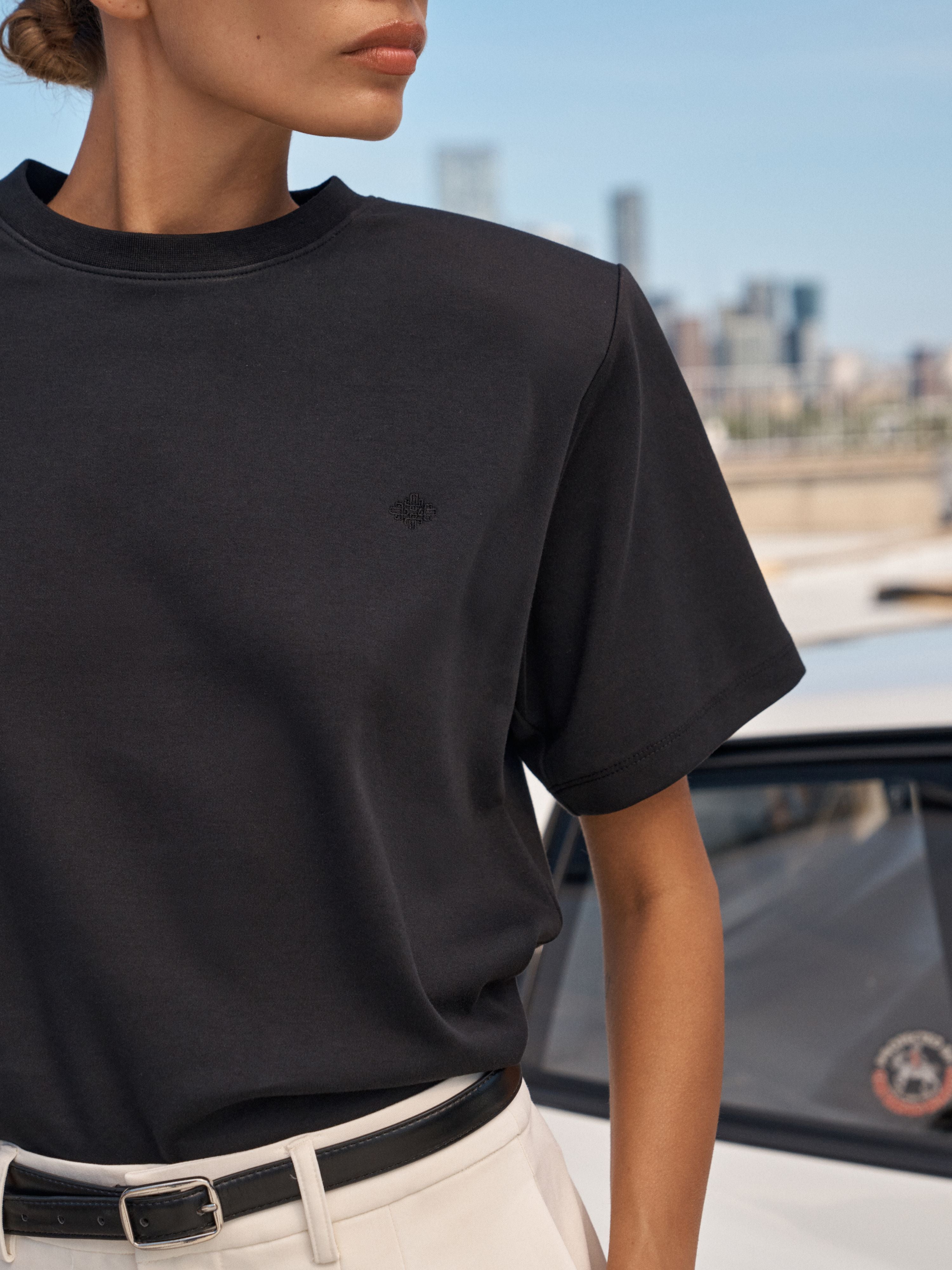 A close-up of a model wearing a black short-sleeve crewneck t-shirt with a small embroidered logo on the chest, paired with white trousers and a black belt, set against a blurred city background.