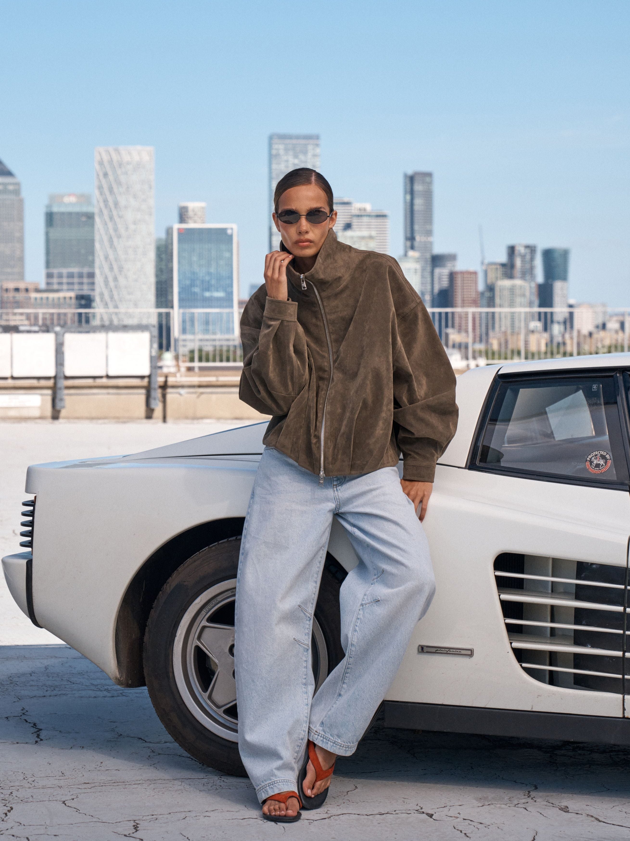 A model wearing a brown suede jacket, light blue wide-leg jeans, and brown sandals leans against a vintage white car with a city skyline in the background.