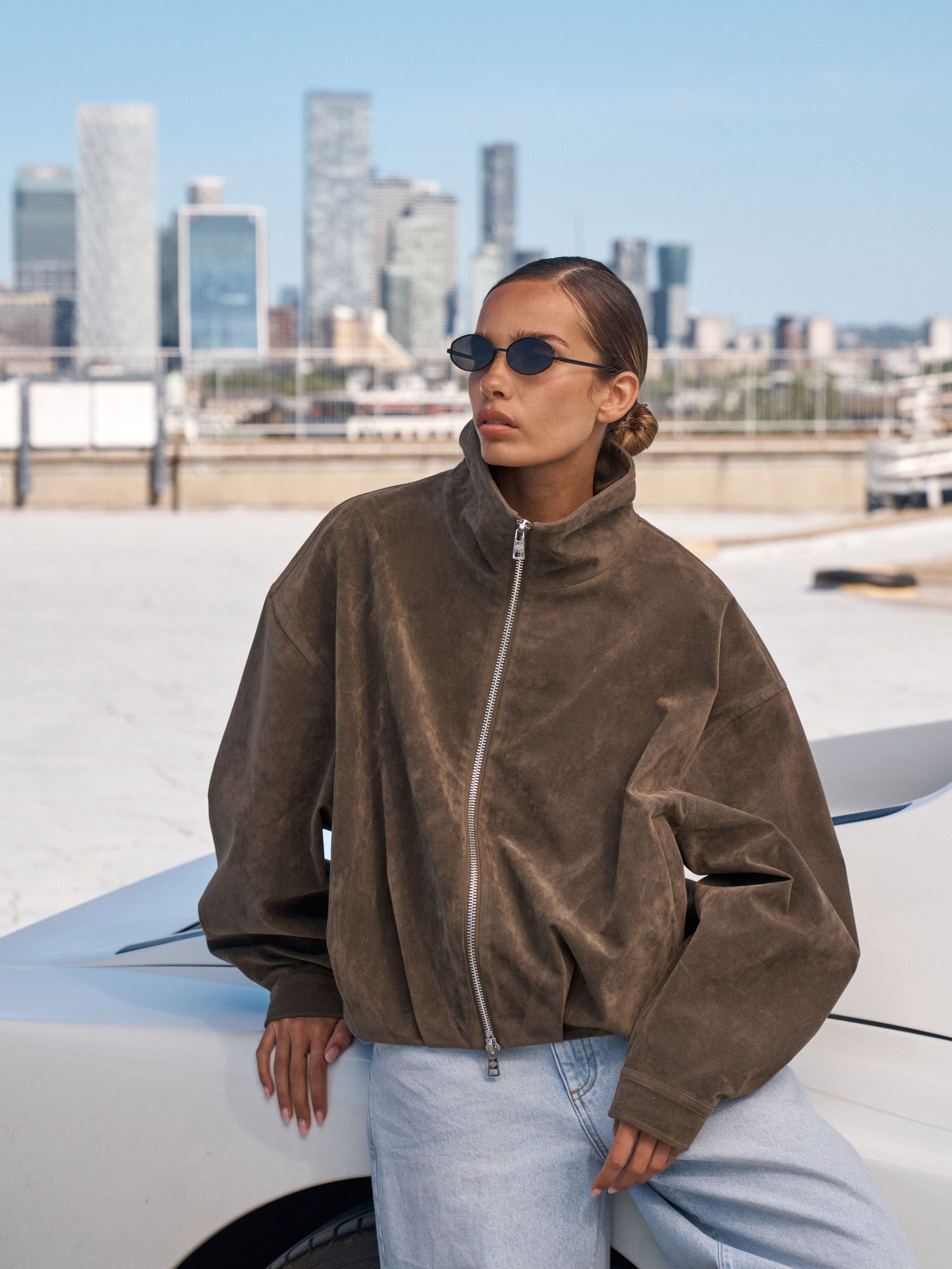 A woman wearing a brown suede zip-up jacket and light blue jeans leans against a white car with a city skyline in the background.