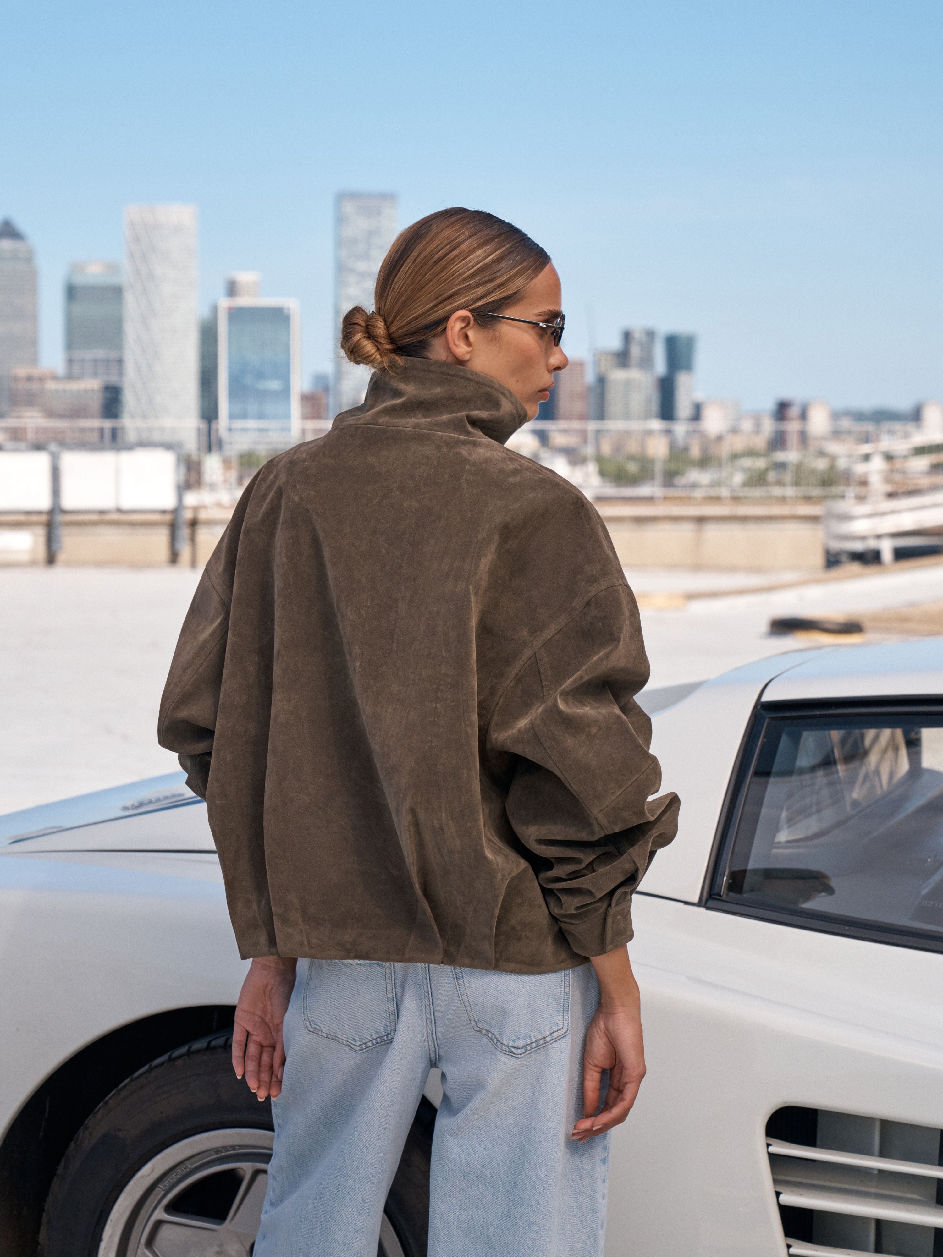 A woman wearing a brown suede jacket and light blue jeans stands on a rooftop next to a white car, with a city skyline in the background under a clear blue sky.
