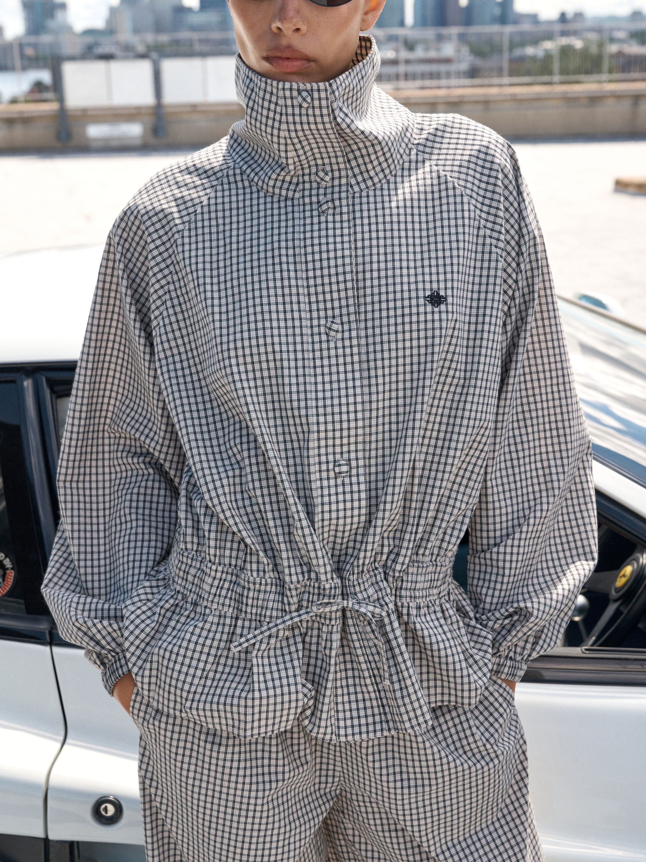 A person wearing a high-neck, long-sleeve button-front shirt and matching pants in a small-scale black and white gingham check pattern, standing outdoors near a white car.