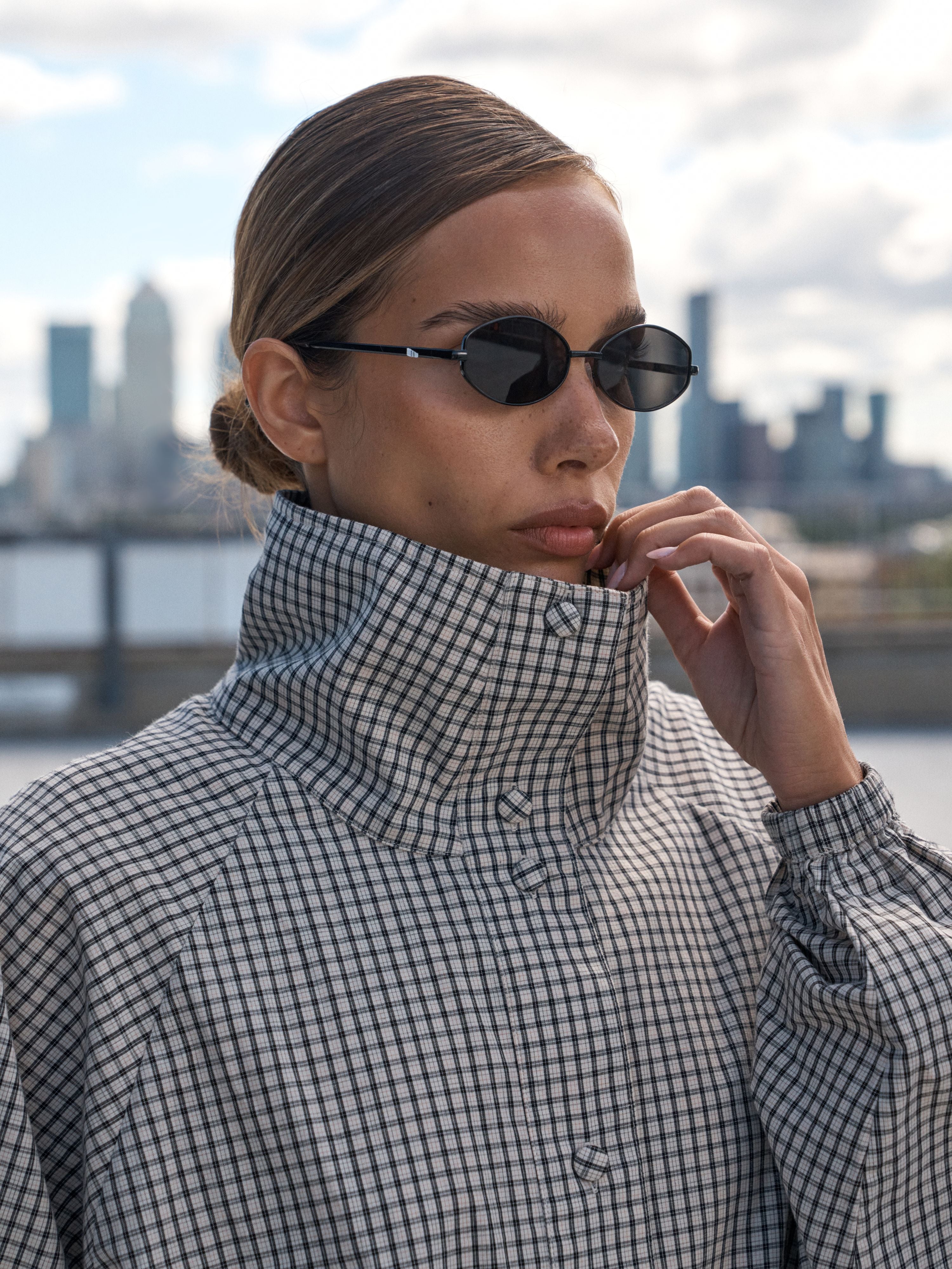 A woman wearing a black and white checkered high-neck button-up shirt and black oval sunglasses, posing outdoors with a blurred city skyline and water in the background.