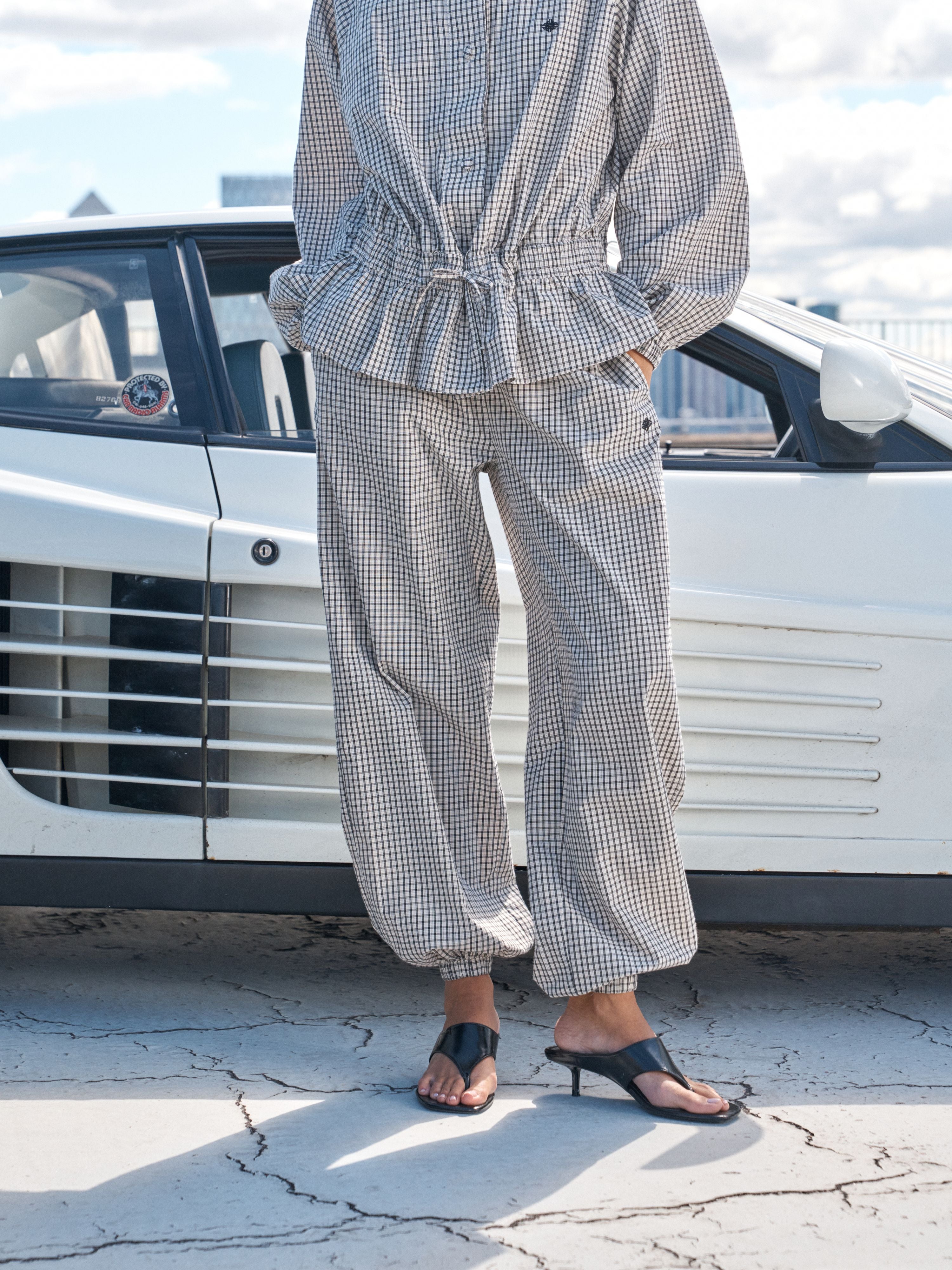 A person wearing a matching two-piece gingham set with a peplum top and wide-leg pants, standing outdoors in front of a white car on a cracked pavement surface.