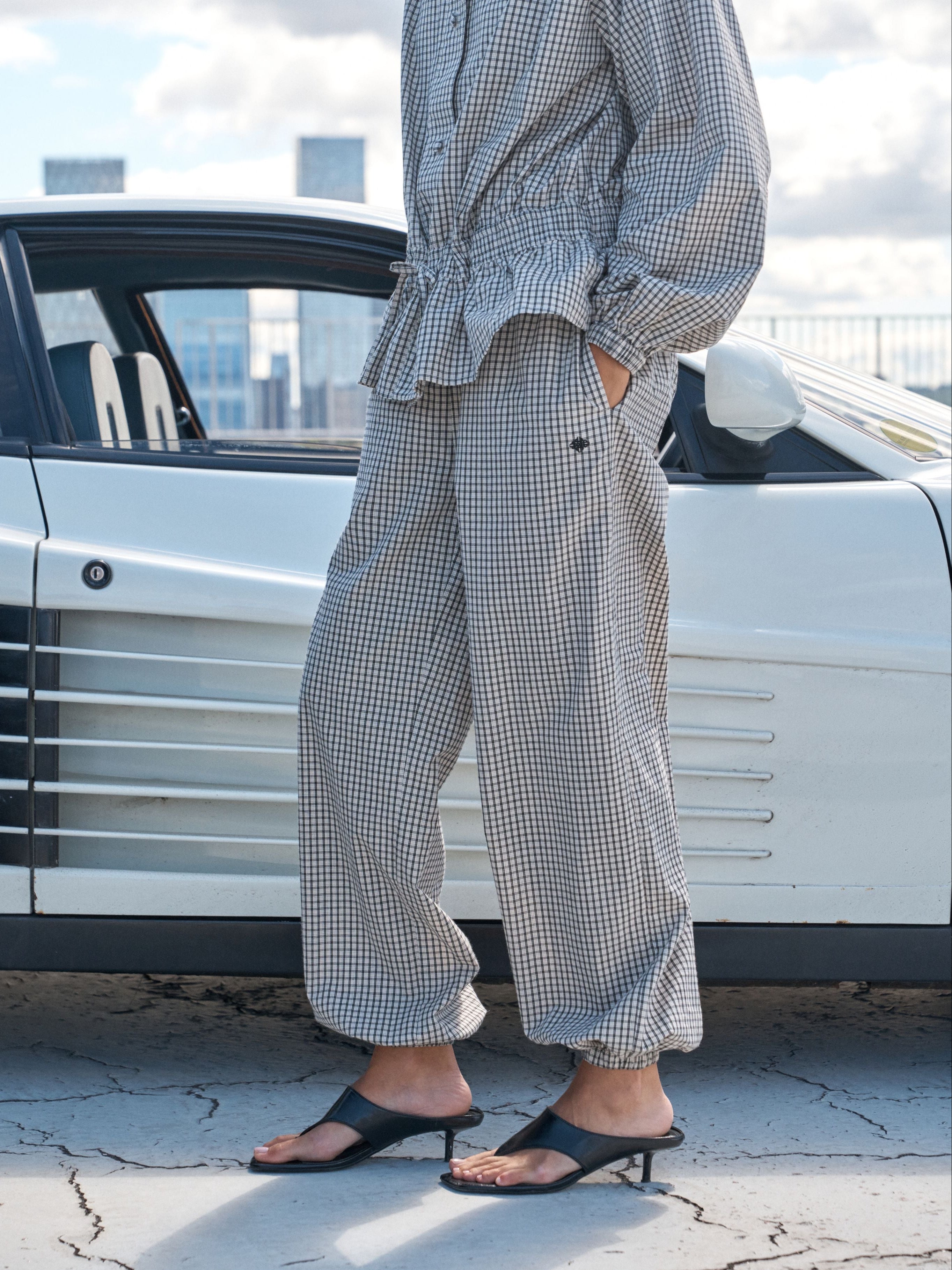 A person wearing a matching beige and black gingham long-sleeve top and wide-leg pants, paired with black slide sandals, standing outdoors next to a white classic car.