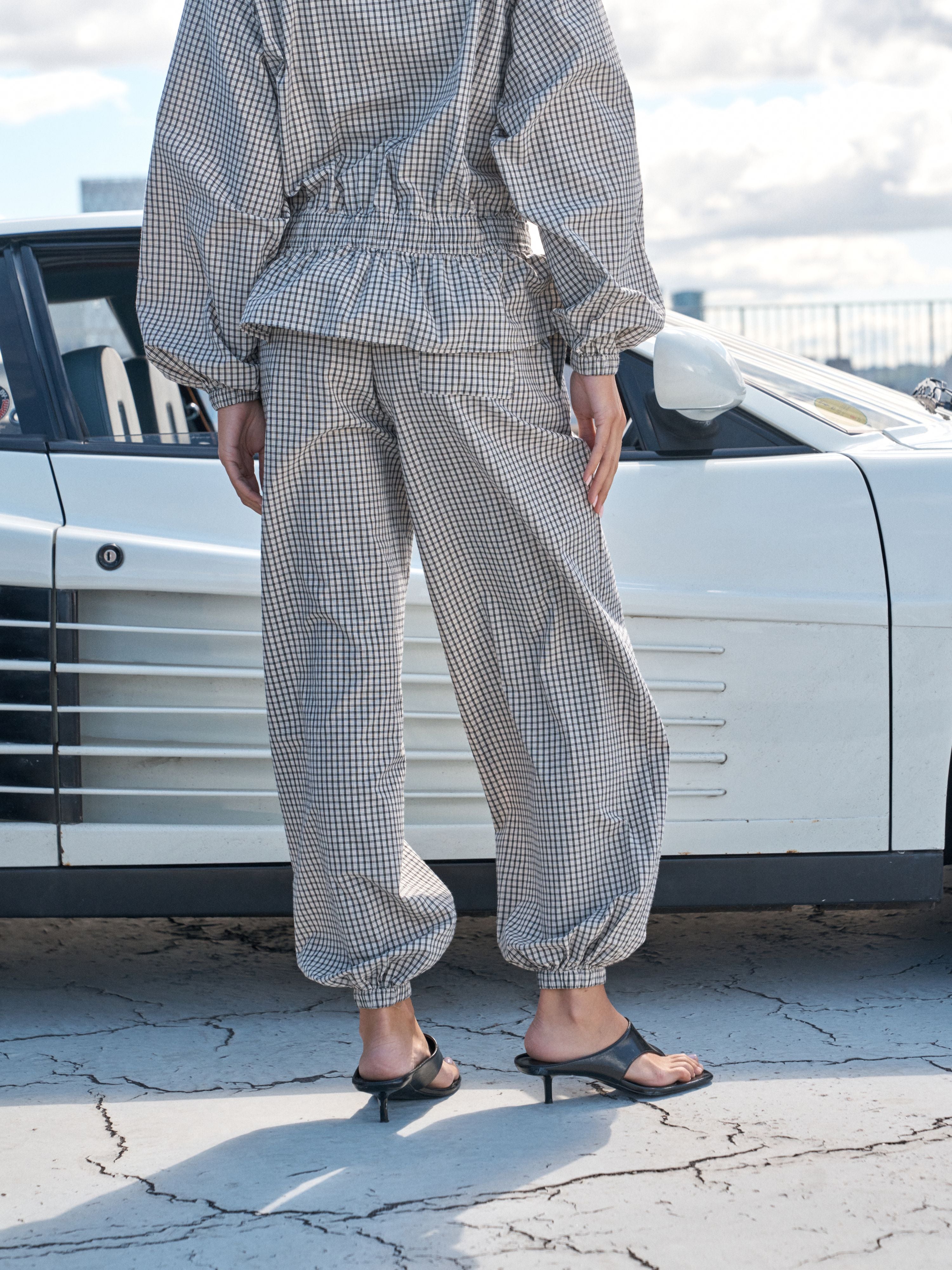A person wearing a matching two-piece gingham outfit with a peplum waist and baggy cropped pants, standing outdoors in front of a white car and wearing black heeled slide sandals.