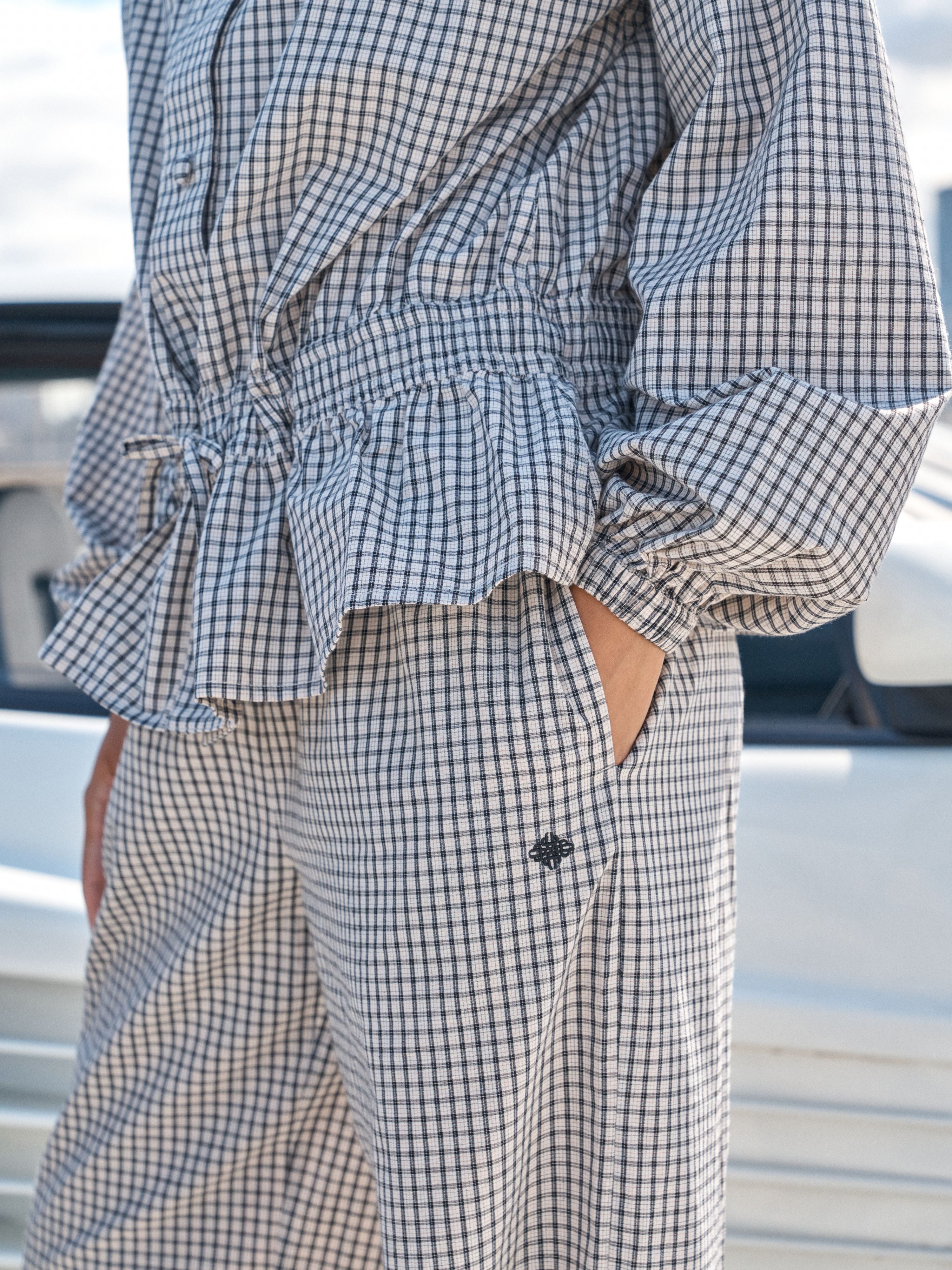 A close-up of a person wearing a blue and white gingham checkered jumpsuit with a ruffled peplum waist, long sleeves, and side buttons.