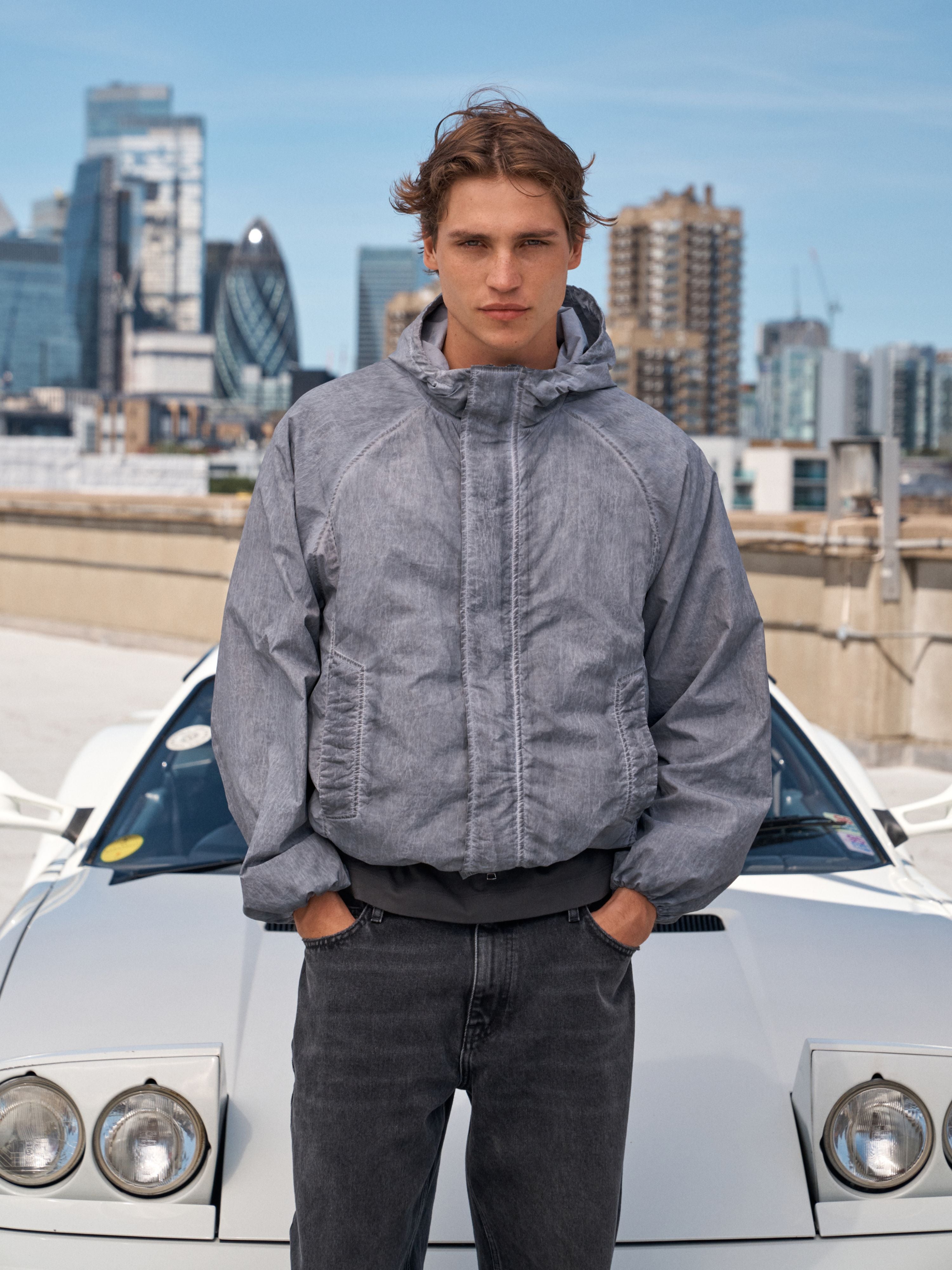 A man wearing a gray hooded puffer jacket and dark gray jeans stands in front of a white sports car with a city skyline in the background.