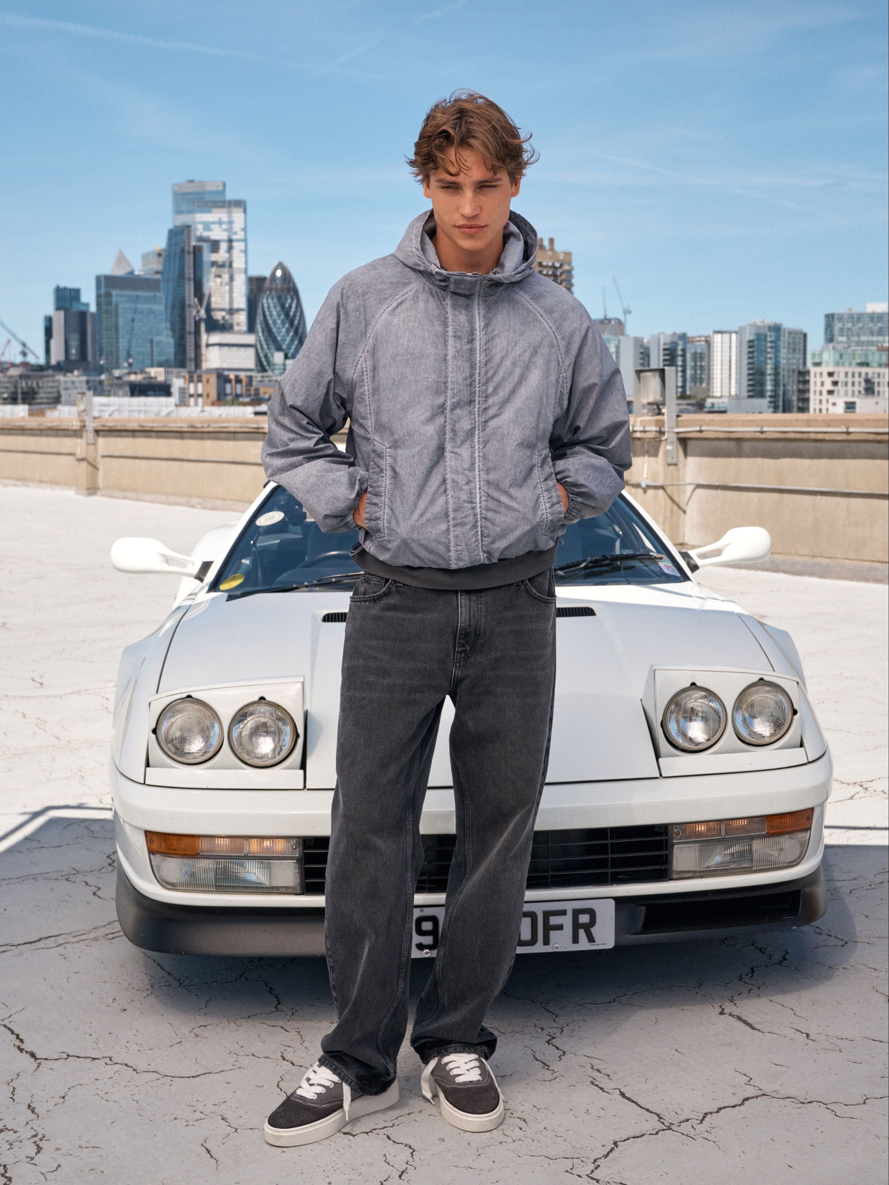 A man wearing a grey zip-up hoodie, dark gray jeans, and black-and-white sneakers stands in front of a white classic sports car on a rooftop with a city skyline in the background.
