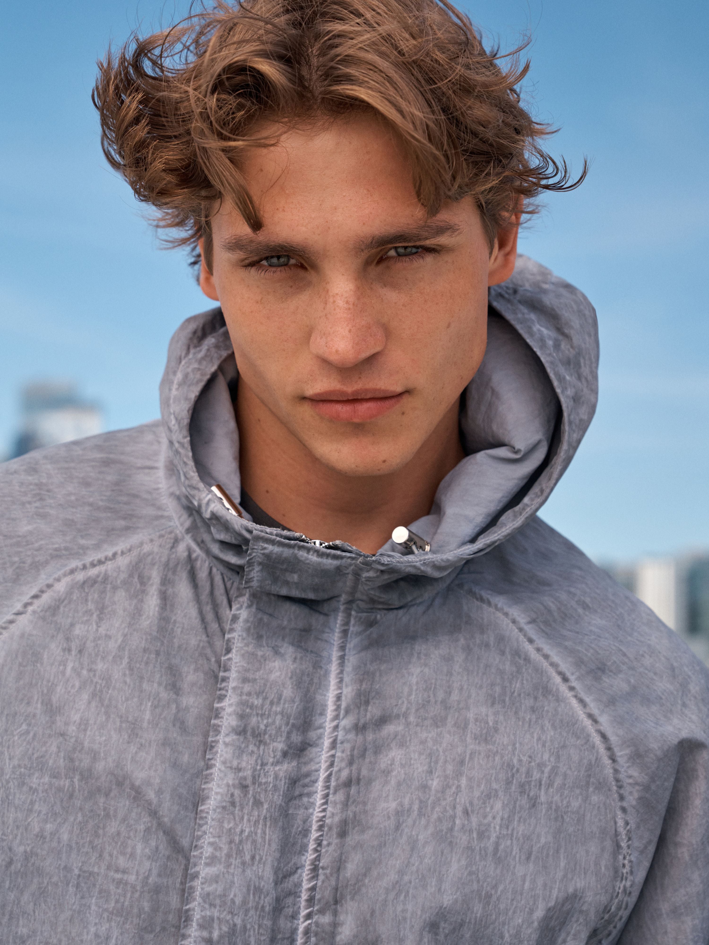 A close-up portrait of a young man wearing a textured gray hooded jacket against a blue sky background.