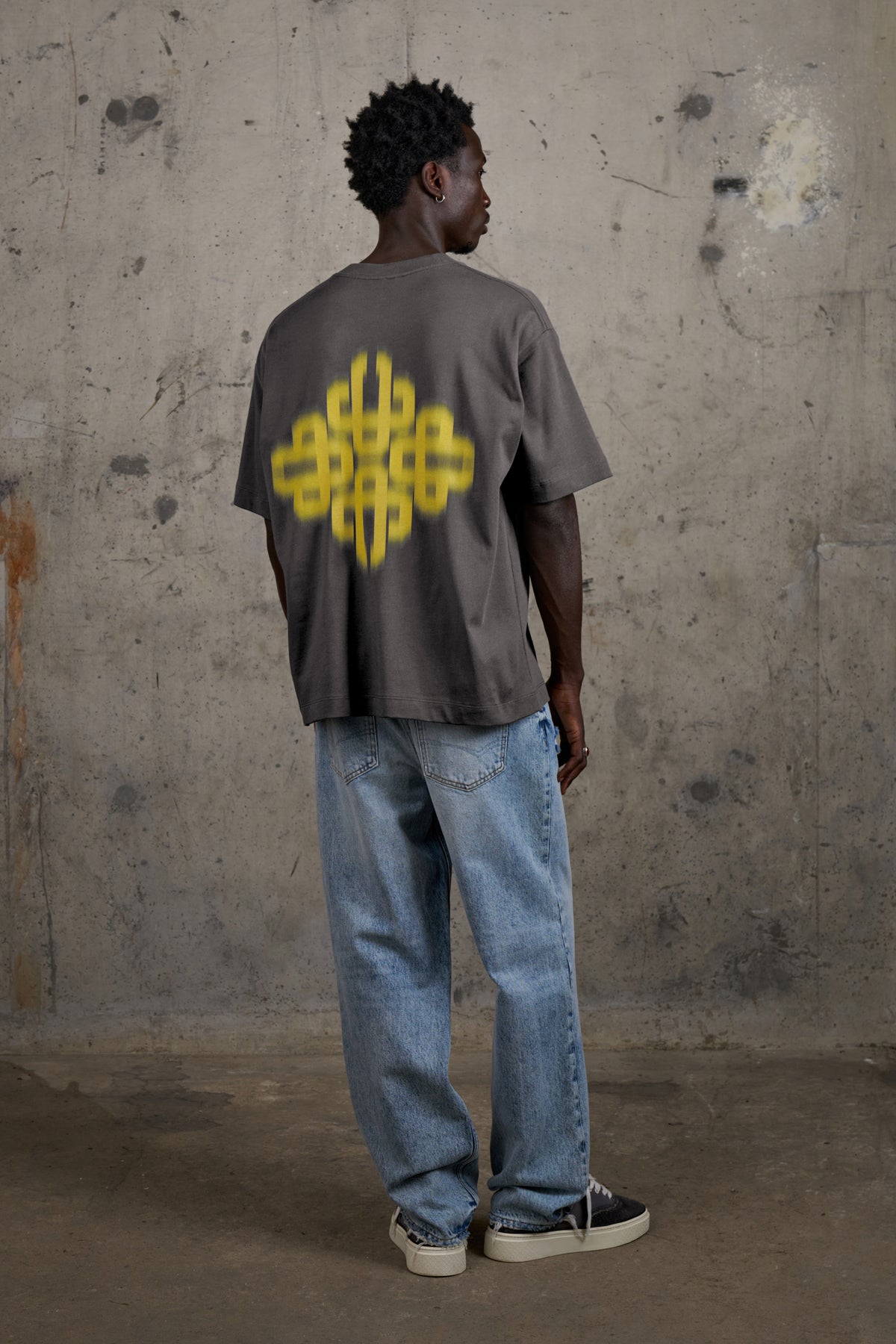 Model wearing the Motion Blurred Emblem T-Shirt in Charcoal/Yellow, full-length back view
