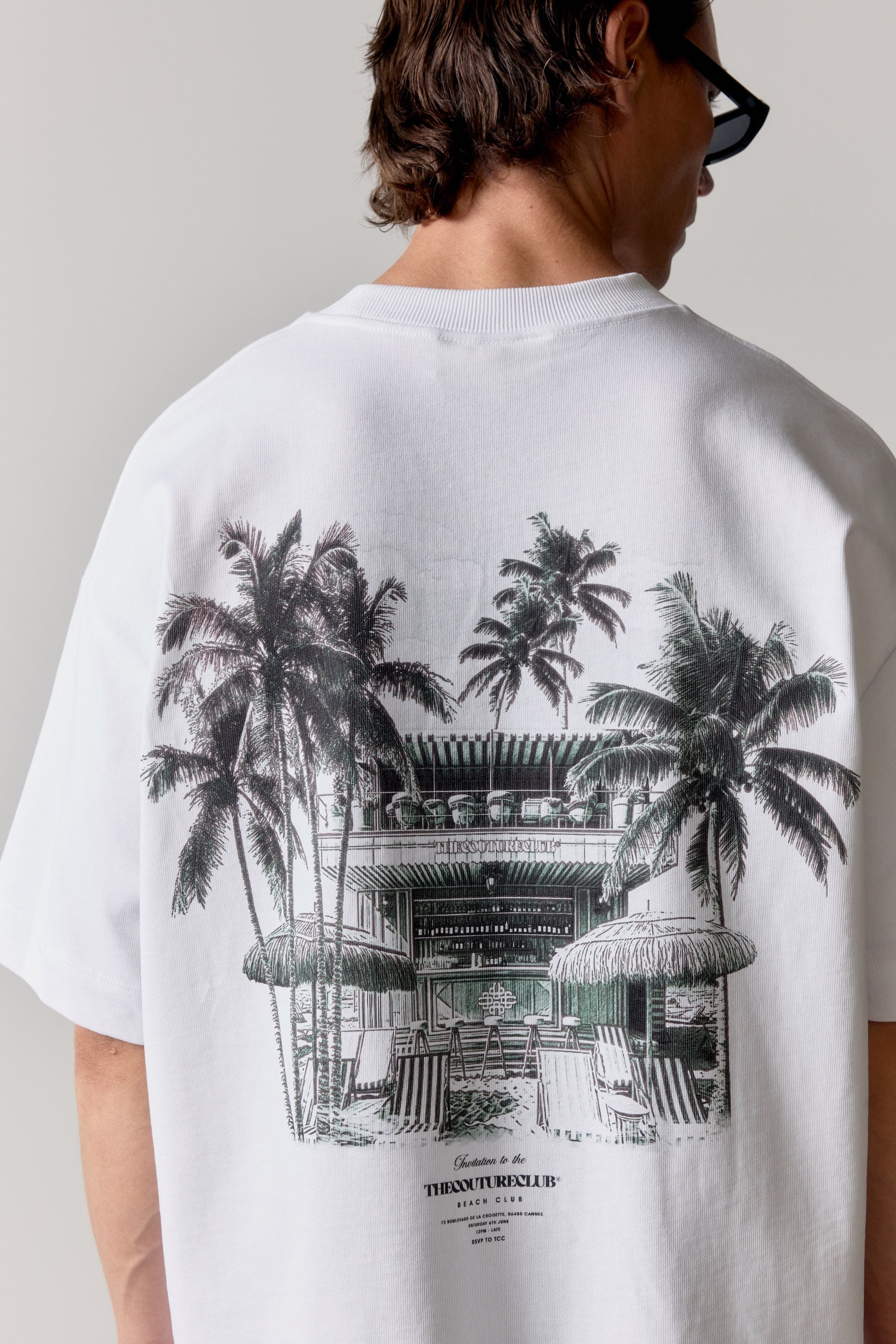 Person wearing a white t-shirt with green graphic of a beach club and palm trees on the back.