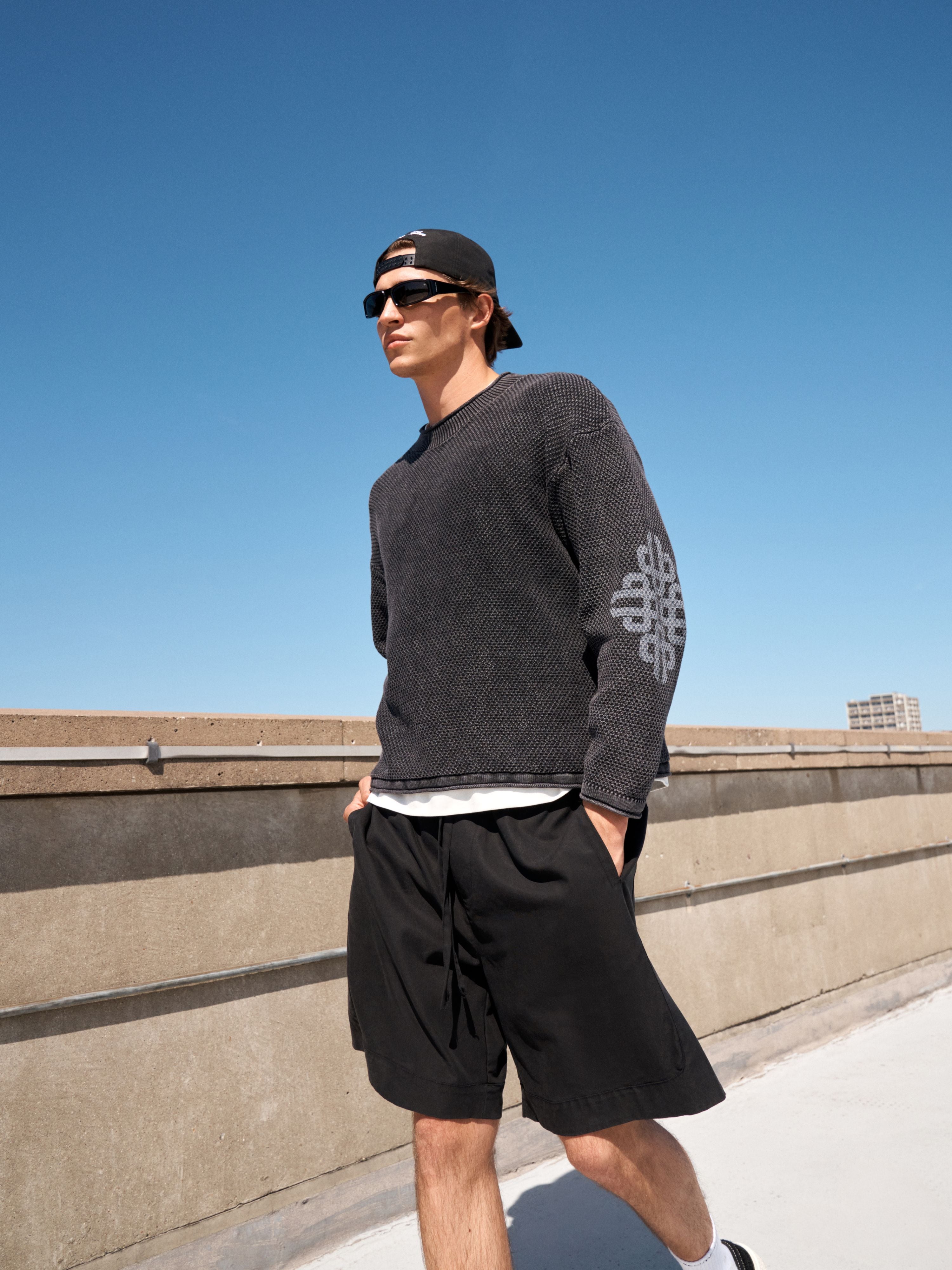 A man wearing a dark gray textured knit sweater with a white geometric pattern on the sleeve, black shorts, a black cap, and sunglasses, standing on a rooftop against a clear blue sky.
