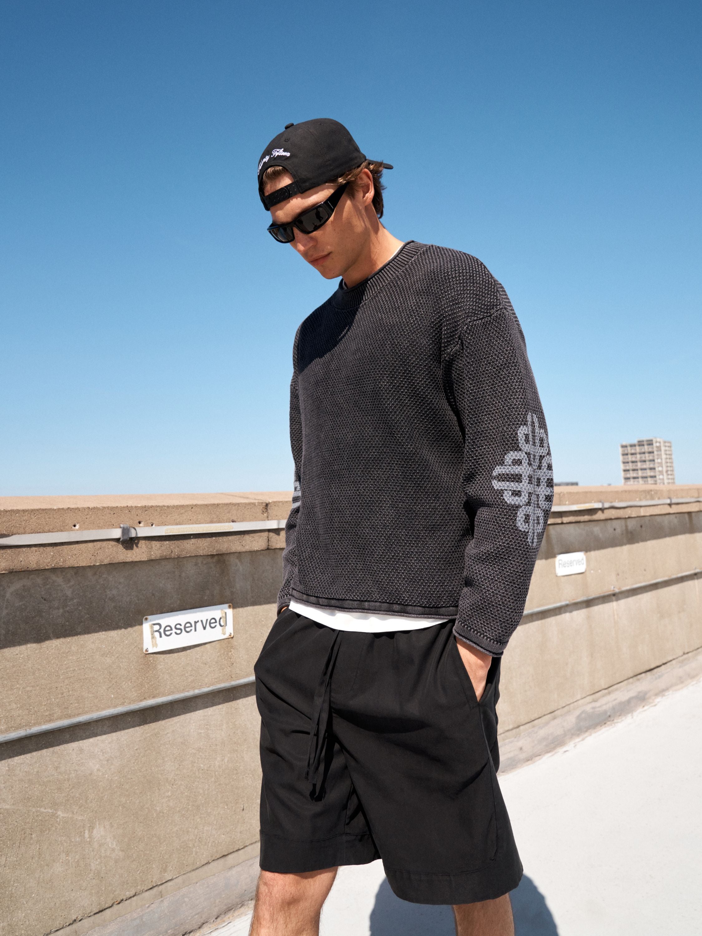 A man wearing a dark gray textured knit sweater with a white geometric pattern on the sleeve, black shorts, a black cap, and sunglasses, standing on a rooftop under a clear blue sky.