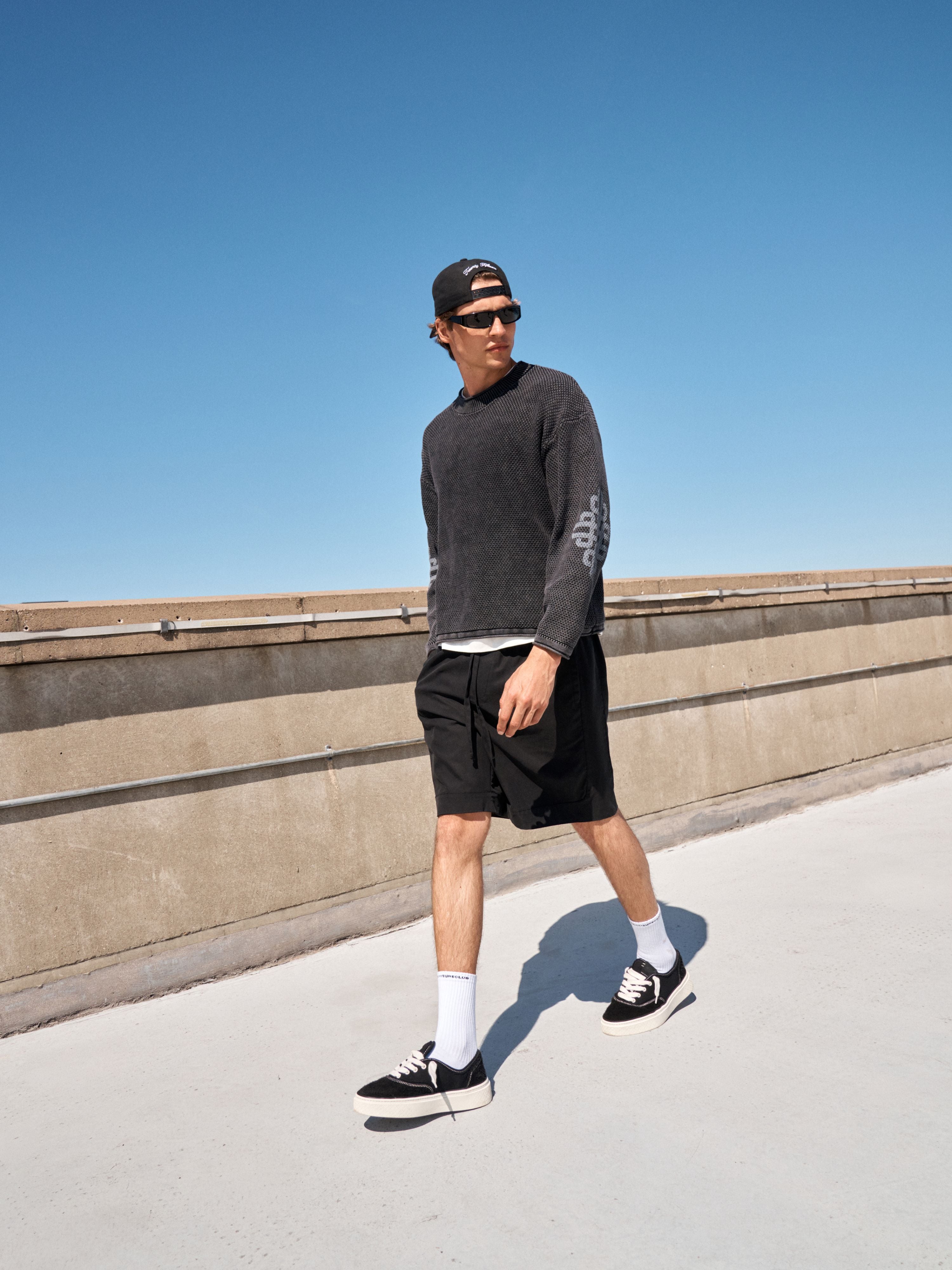 A man walking outdoors on a sunny day wearing a dark charcoal gray long-sleeve crewneck sweatshirt, black cargo shorts, white crew socks, black sneakers with white soles, and a black baseball cap.