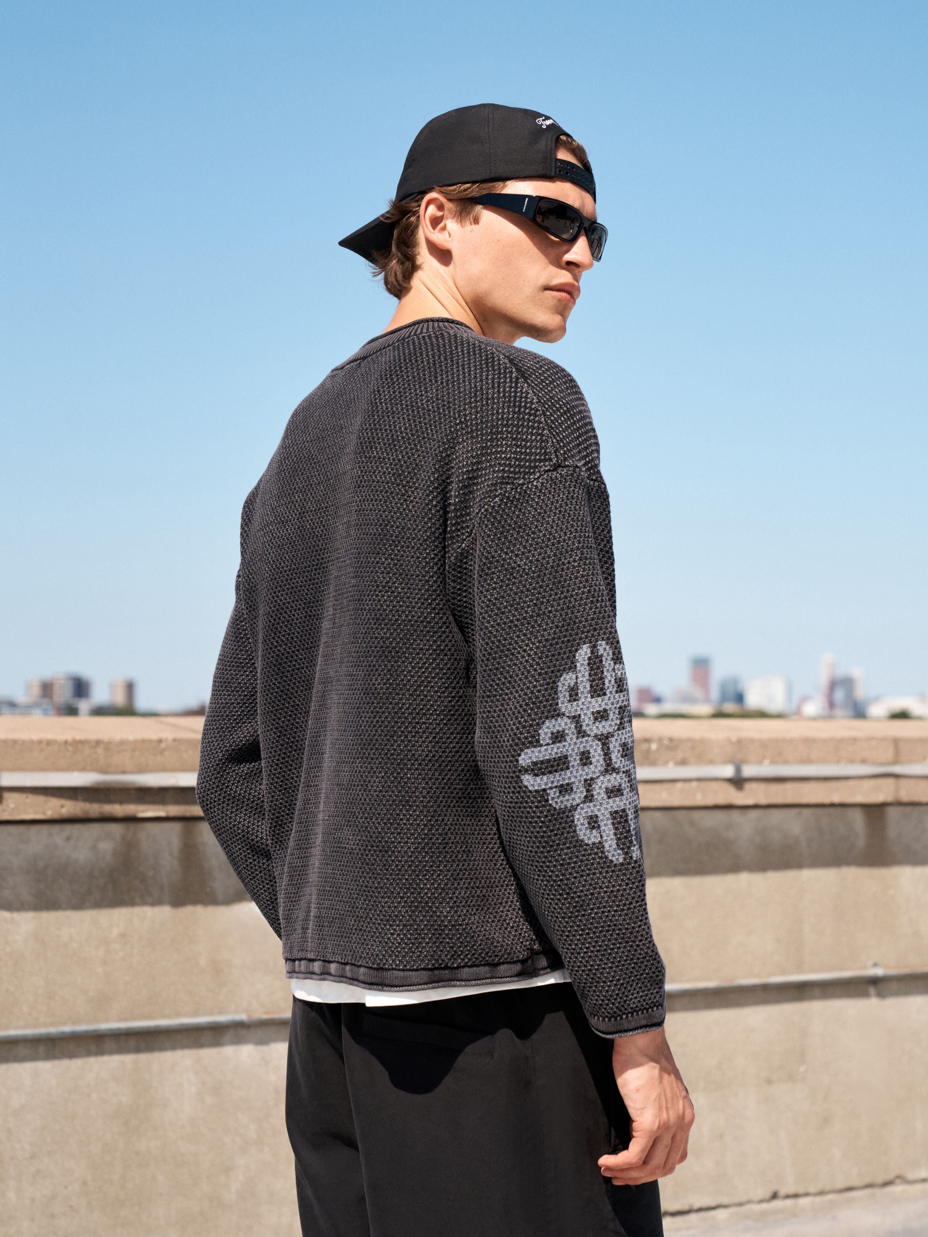 A man wearing a dark gray waffle-knit sweater with a white geometric pattern on the sleeve, black pants, a black cap, and sunglasses, standing outdoors on a concrete structure with a city skyline and blue sky in the background.