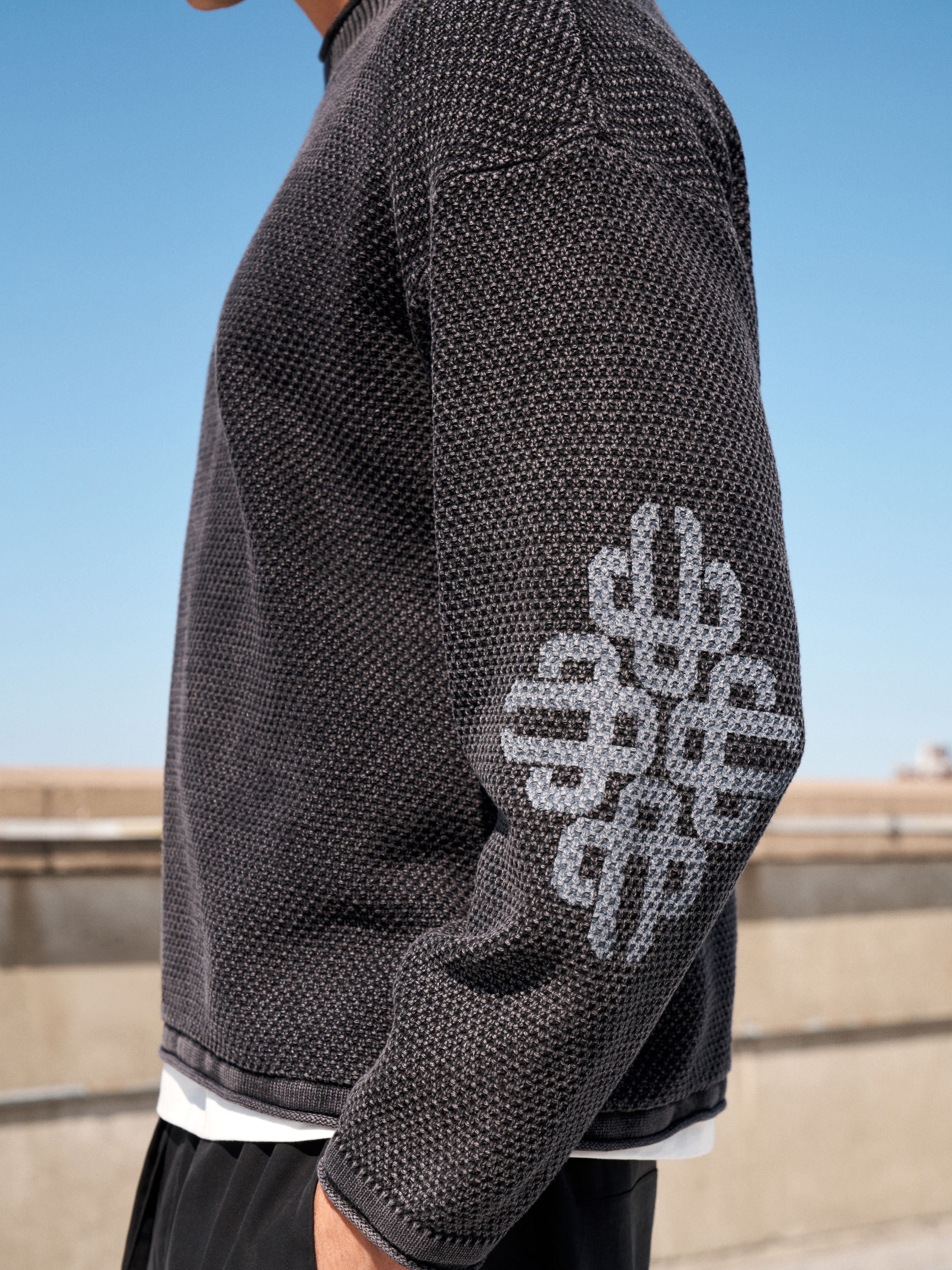 A close-up side view of a person wearing a dark charcoal gray textured knit sweater with a large light gray geometric logo pattern on the sleeve, set against an outdoor background with a clear blue sky.