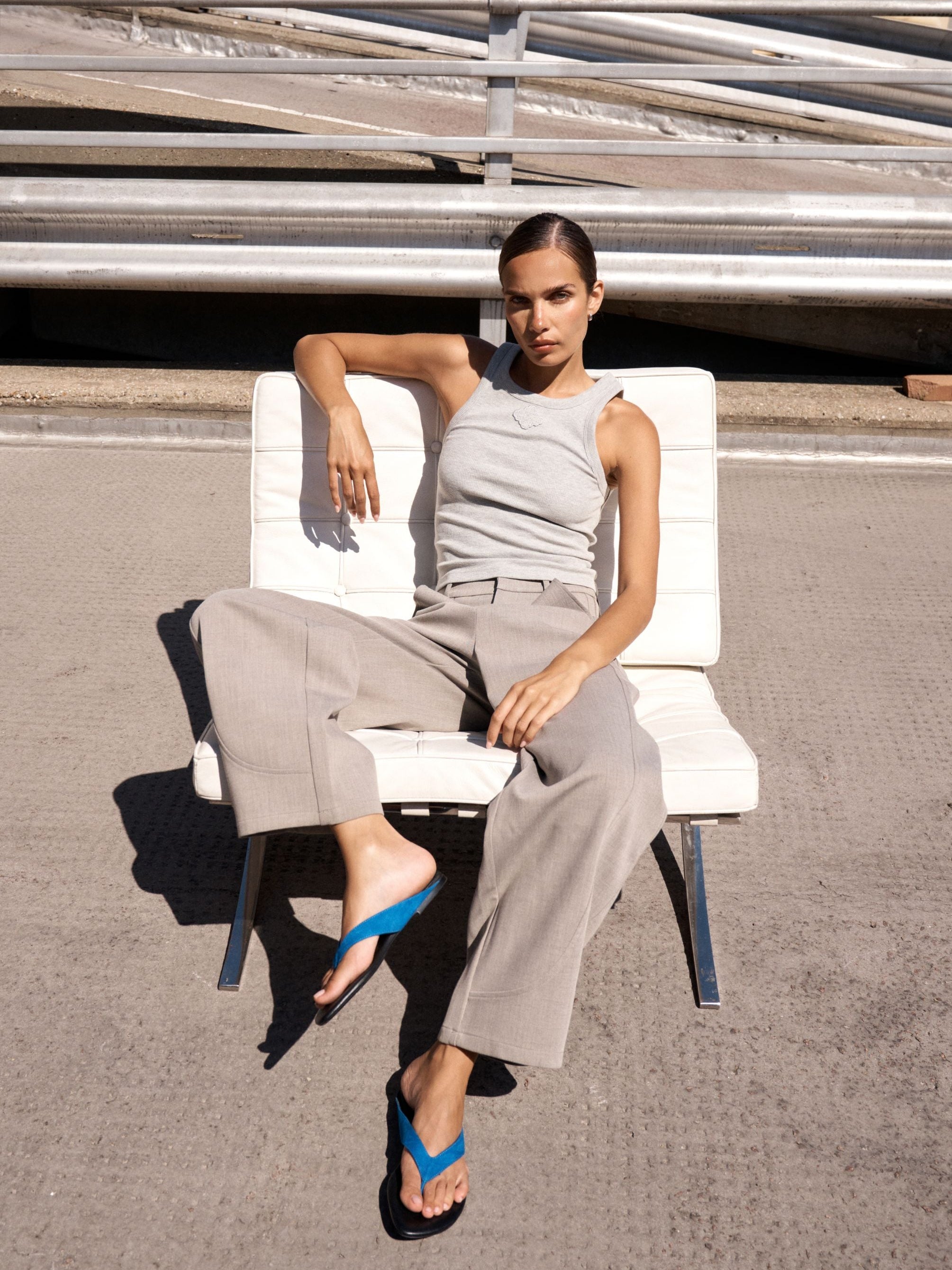 A woman sits on a white lounge chair outdoors on a paved surface, wearing a light gray sleeveless top and matching wide-leg pants, with bright blue flip-flop sandals.