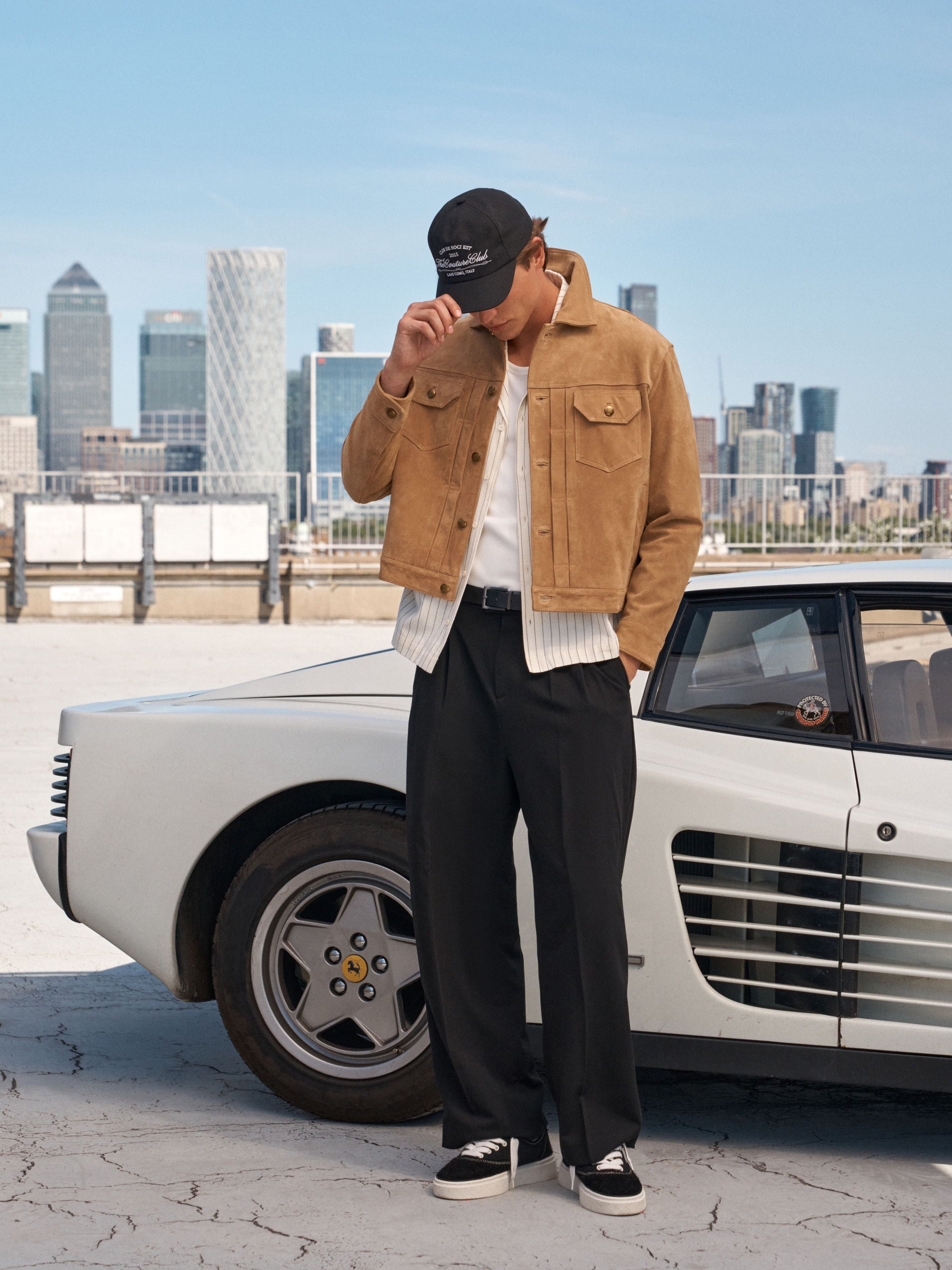 A man stands next to a white vintage car in an outdoor urban setting, wearing a tan jacket, white shirt, black trousers, black sneakers, and a black cap with a white logo.
