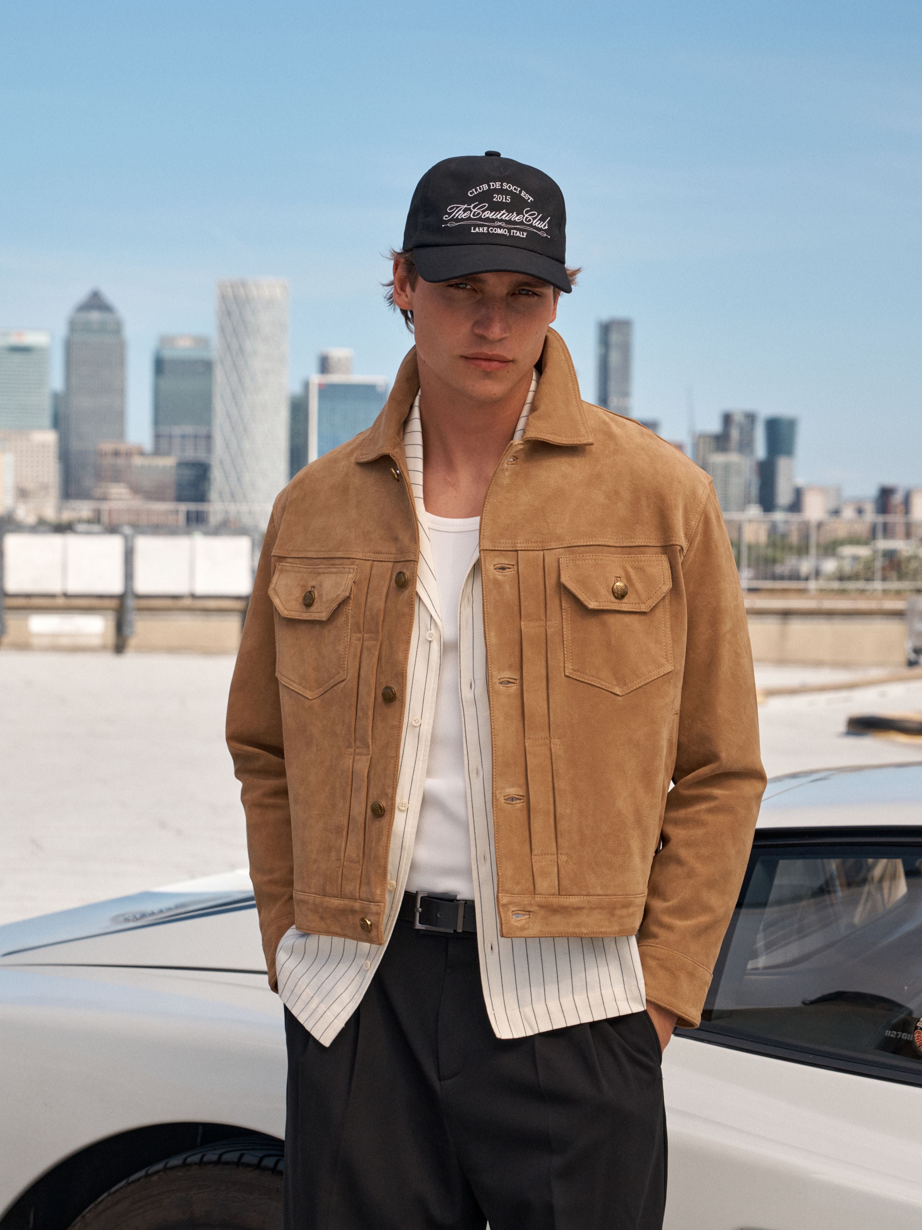 A man wearing a tan suede jacket over a white striped shirt and black trousers, standing outdoors with a city skyline and a white car in the background.