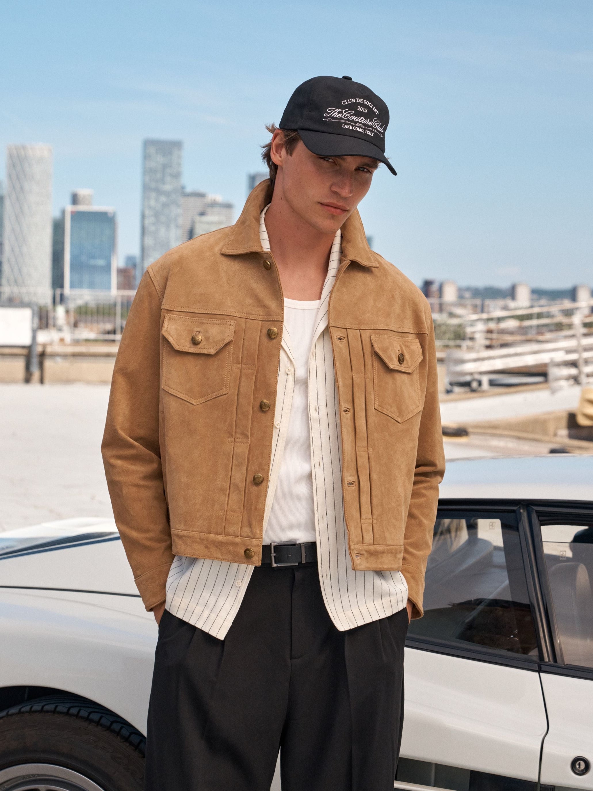 A man wearing a tan suede jacket, white striped shirt, black trousers, and a black baseball cap stands outdoors next to a white car with a city skyline in the background.