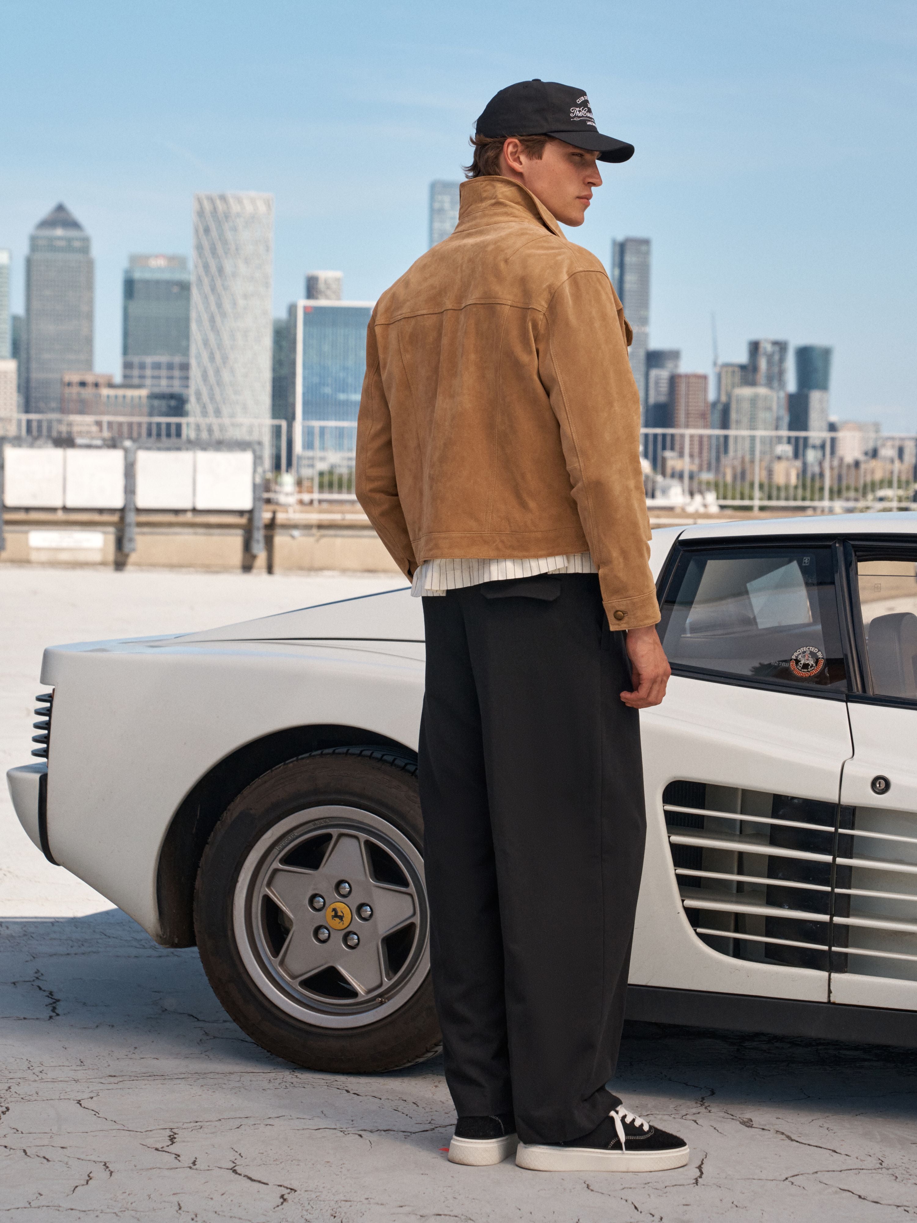A man wearing a tan suede jacket, black trousers, a black cap, and black sneakers stands next to a white classic car with a city skyline in the background.