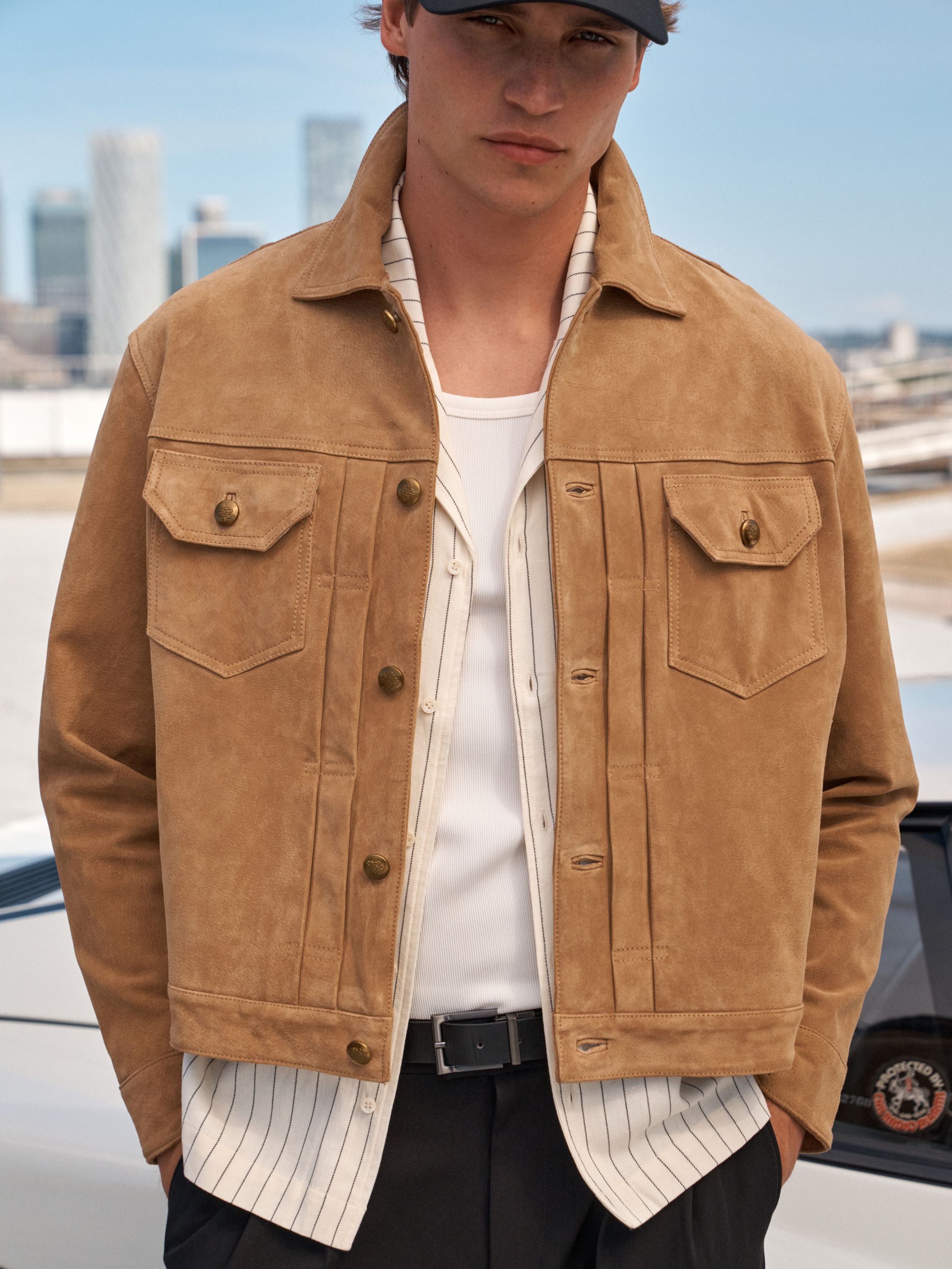 A man wearing a tan suede jacket over a white striped shirt and black trousers, standing outdoors with a blurred cityscape background.