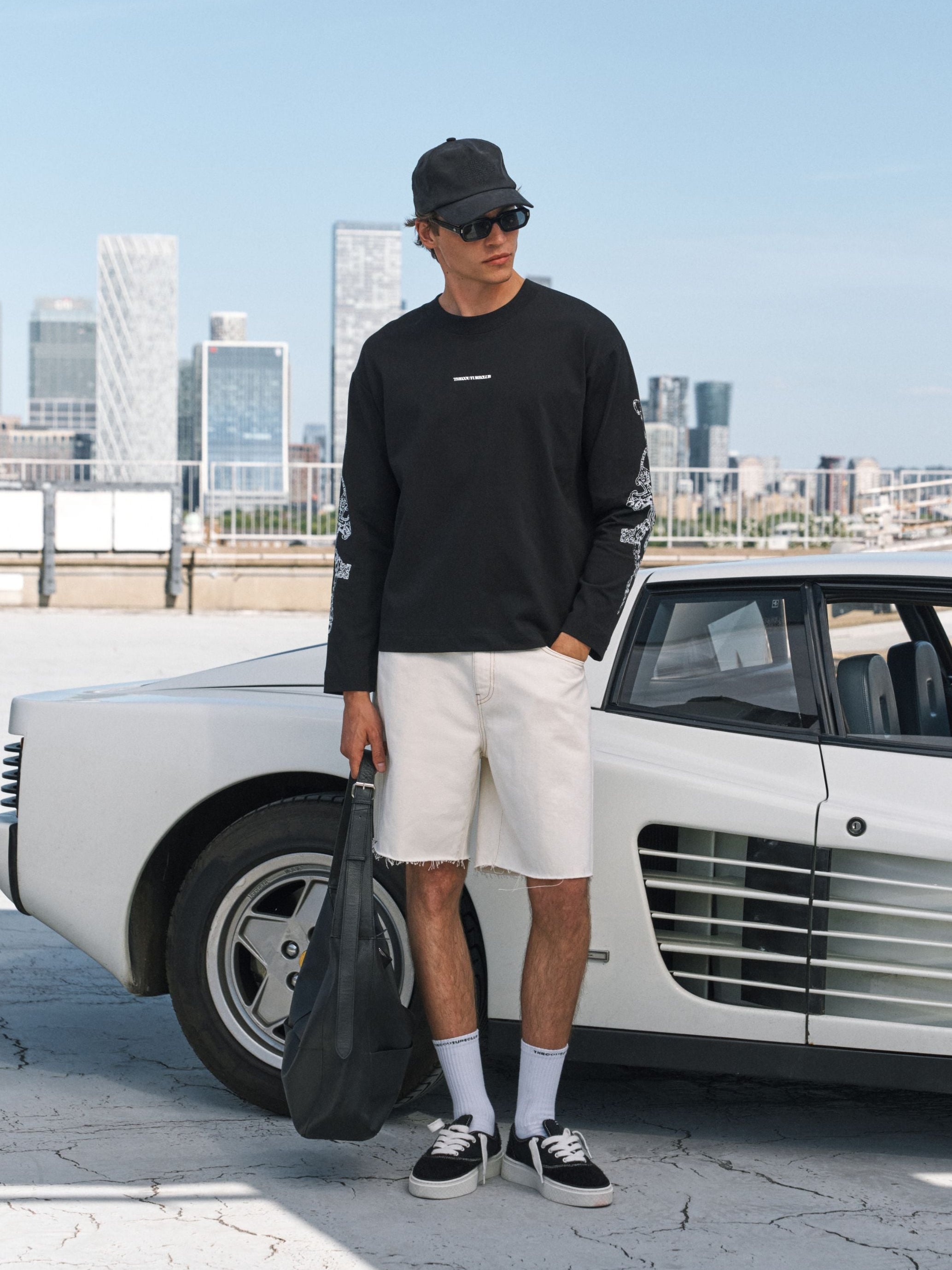 A man wearing a black long-sleeve t-shirt sweatshirt, off-white shorts, white socks, and black sneakers stands next to a white classic sports car, holding a black duffel bag. The background shows a city skyline under a clear blue sky.