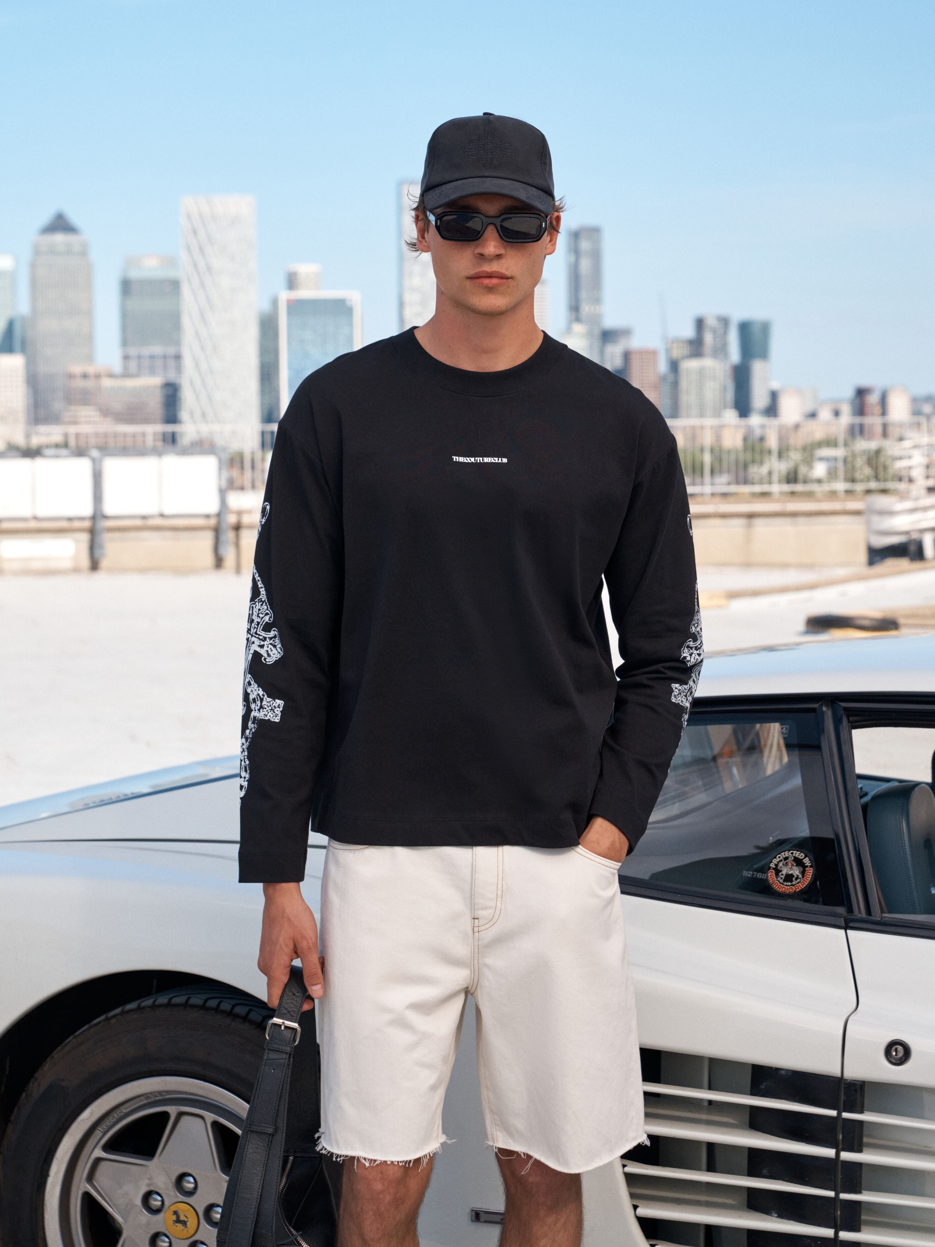 A man wearing a black long-sleeve crewneck t-shirt with white graphic prints on the sleeves, paired with off-white shorts, a black cap, and sunglasses, standing next to a white classic car with a city skyline in the background.