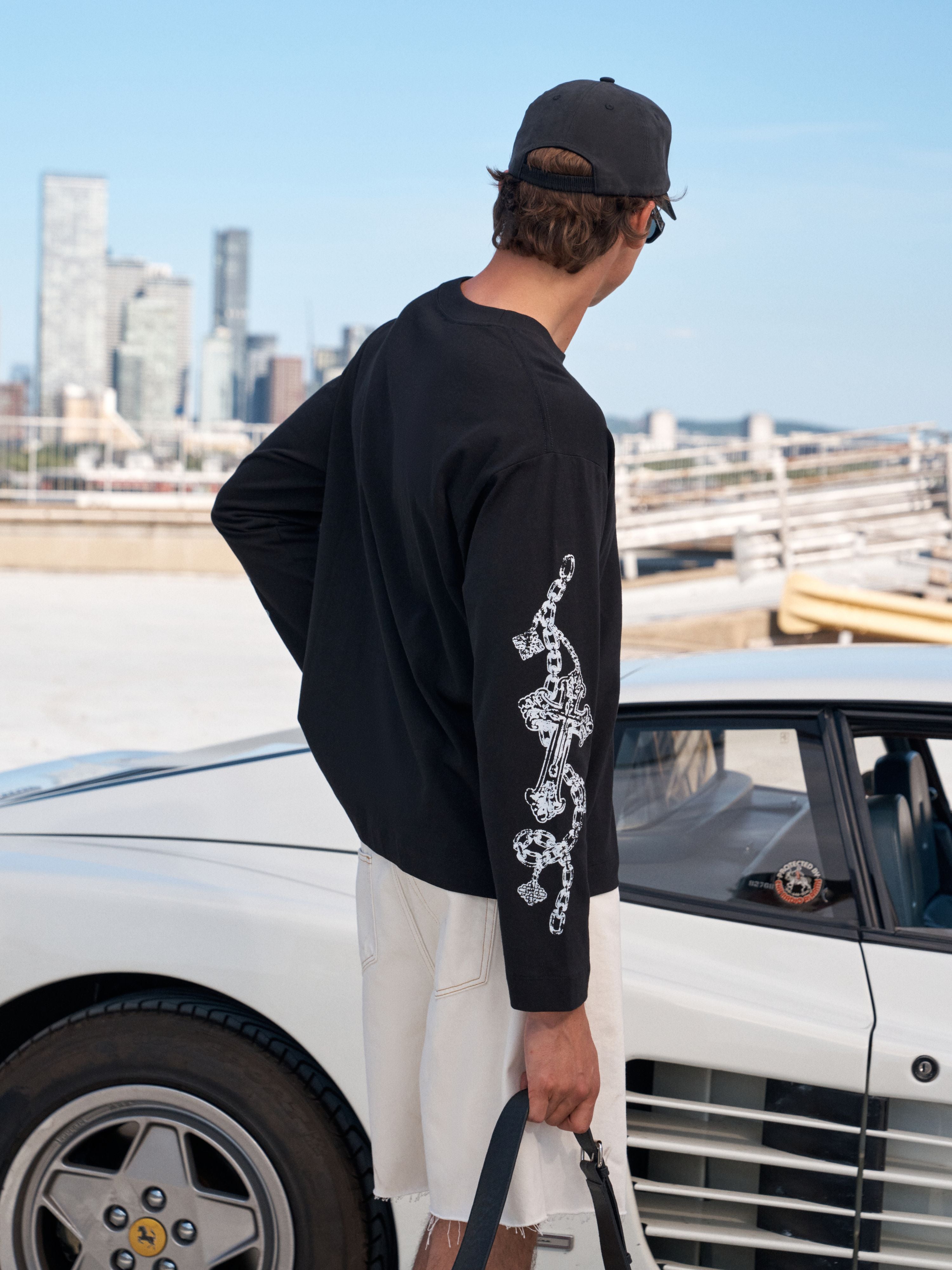 A man wearing a black long-sleeve t-shirt with a white graphic print on the sleeve, paired with white shorts and a black cap, standing next to a white classic car with a city skyline in the background.