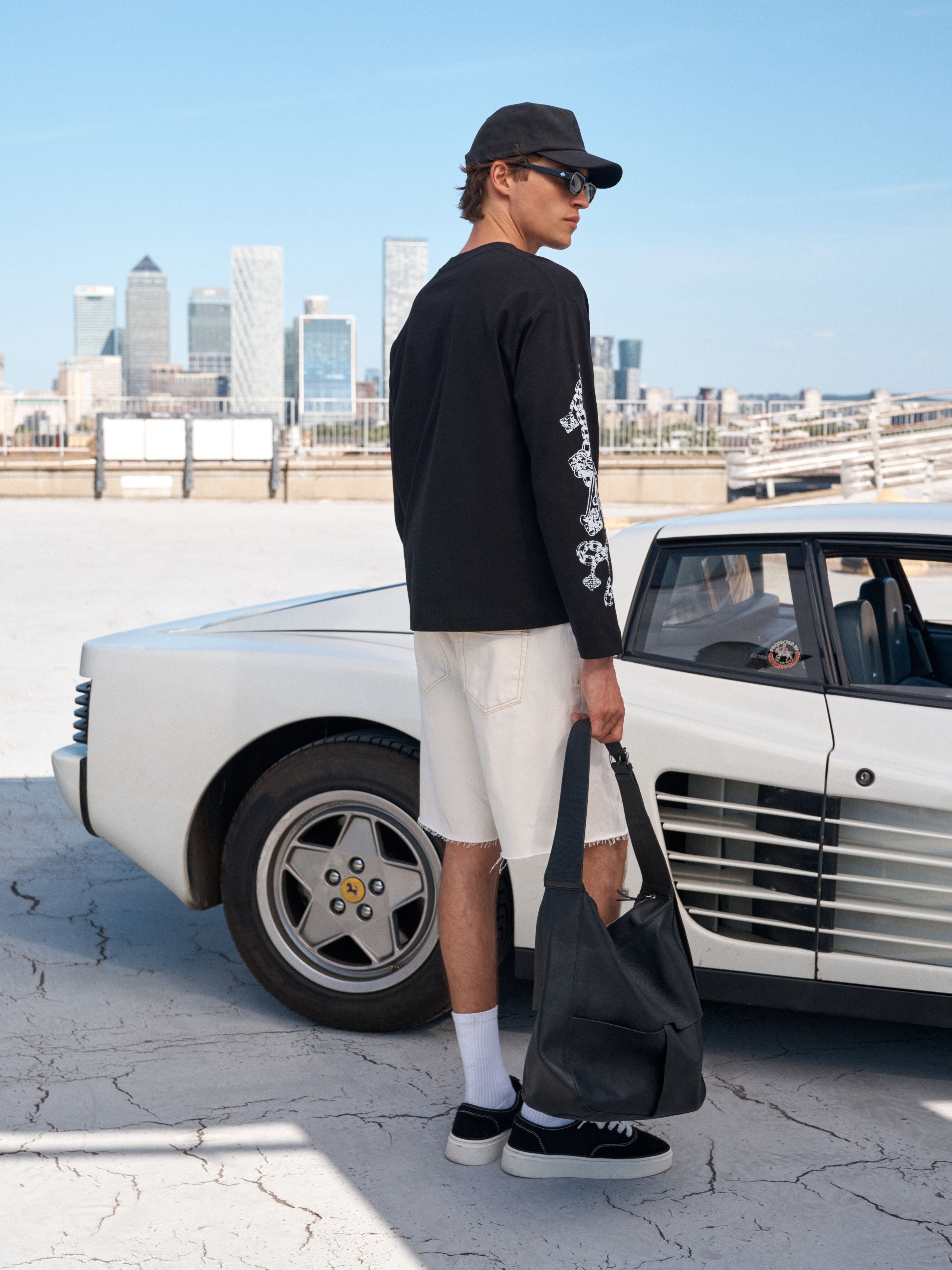 A man wearing a black long-sleeve graphic t-shirt, beige shorts, black sneakers, and a black cap stands next to a white vintage car, holding a black tote bag in an outdoor rooftop setting with a city skyline in the background.