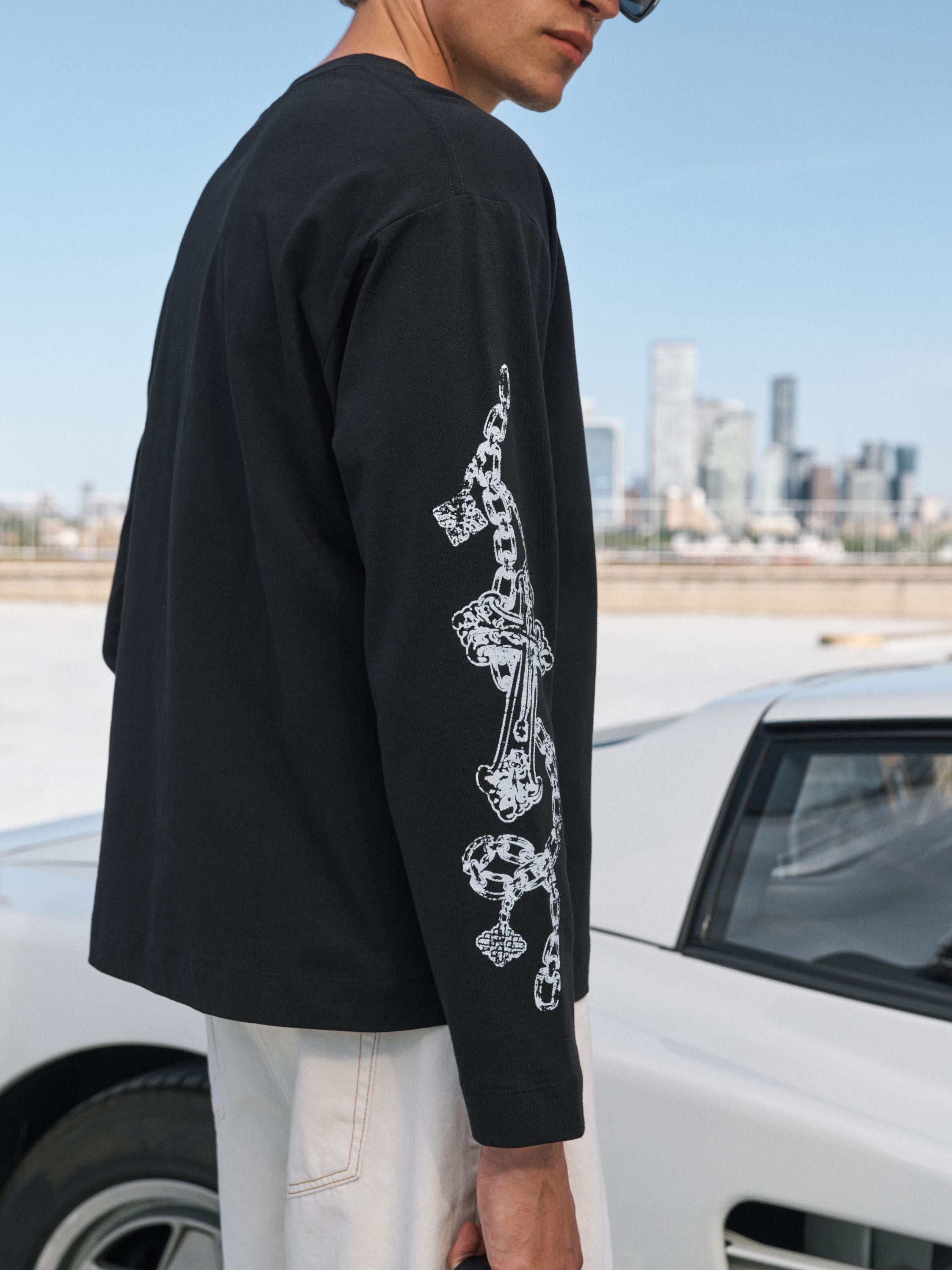 A man wearing a black long-sleeve t-shirt with a white graphic print of a dagger and skull on the sleeve, standing outdoors next to a white car with a city skyline in the background.