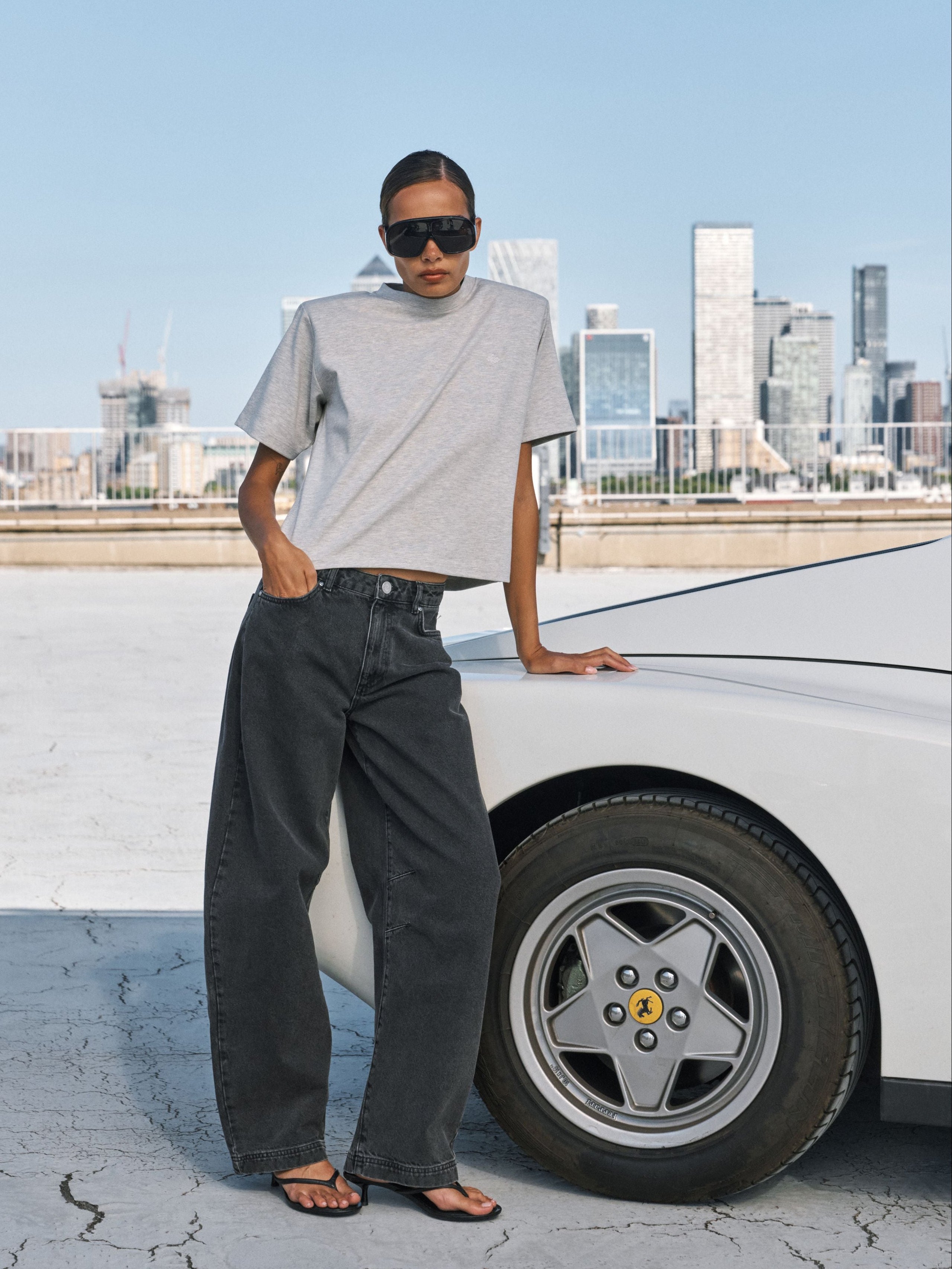 A model wearing a light gray cropped t-shirt and dark gray wide-leg jeans stands next to a white car on a rooftop with a city skyline in the background.