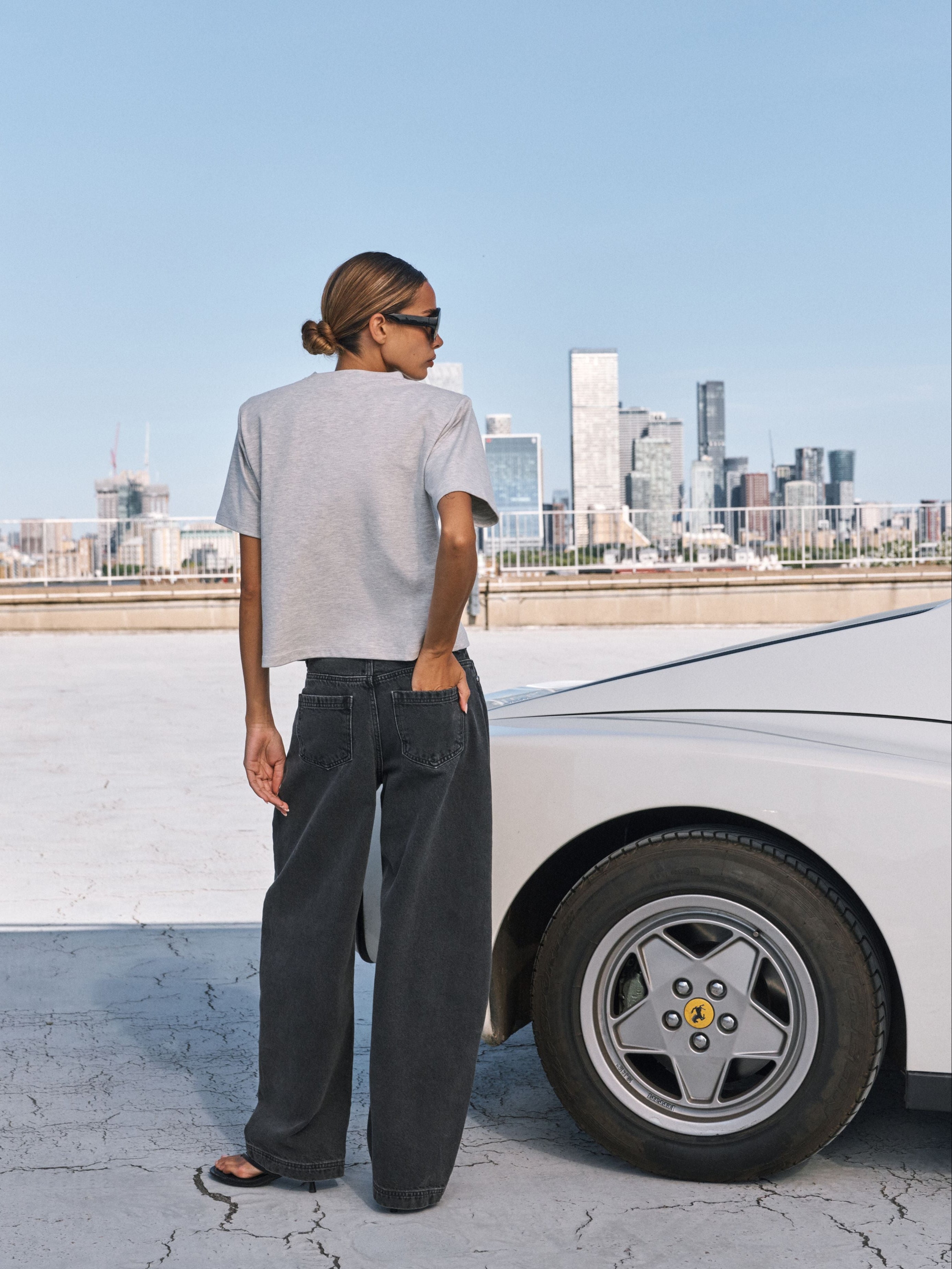 A woman stands with her back to the camera wearing a light gray t-shirt, dark wide-leg jeans, and black sandals, standing next to a white car with a city skyline in the background.