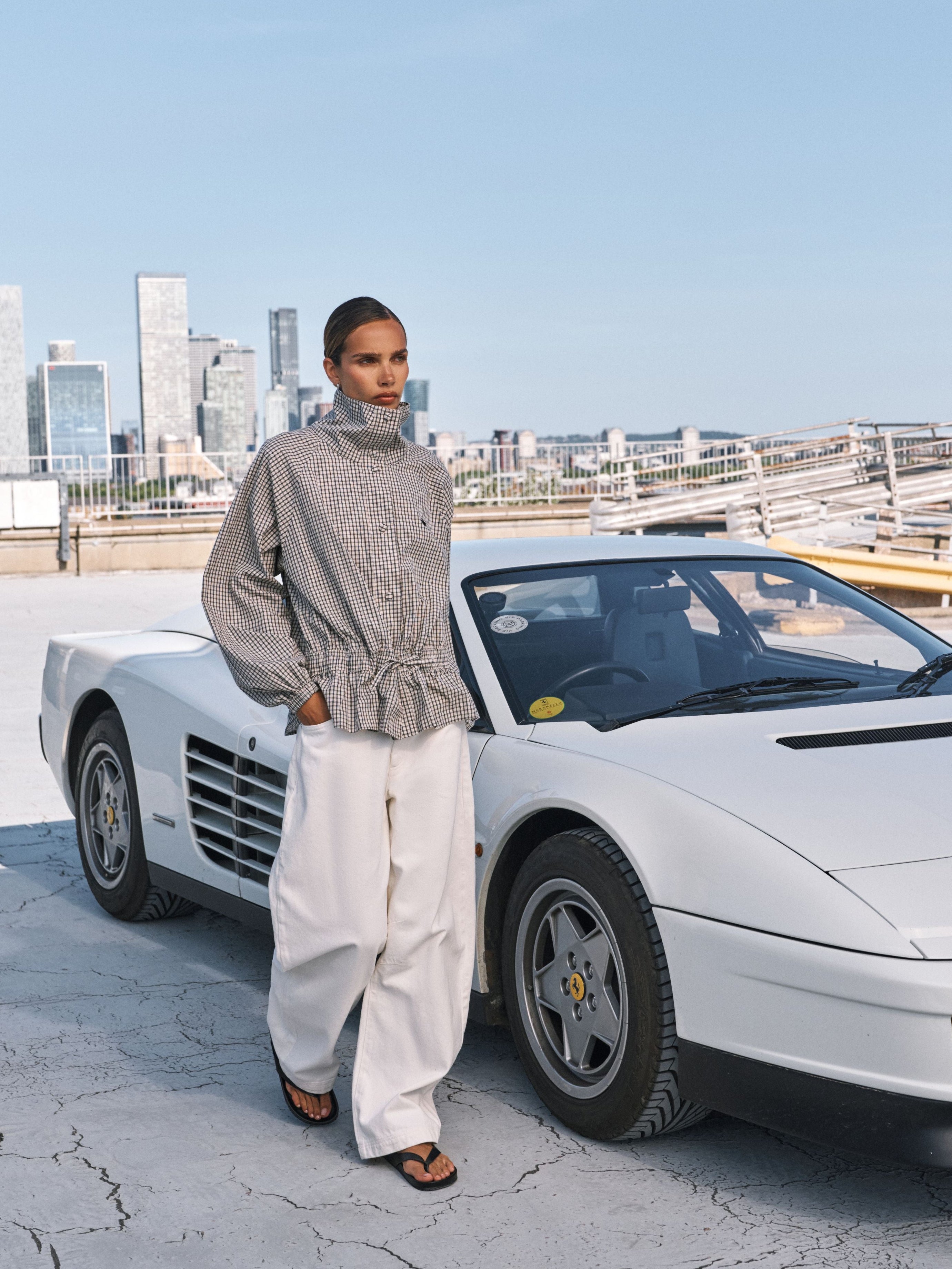A model stands next to a white Ferrari Testarossa on a rooftop with a city skyline in the background. The model is wearing a gray ribbed knit mock-neck top and white wide-leg trousers.
