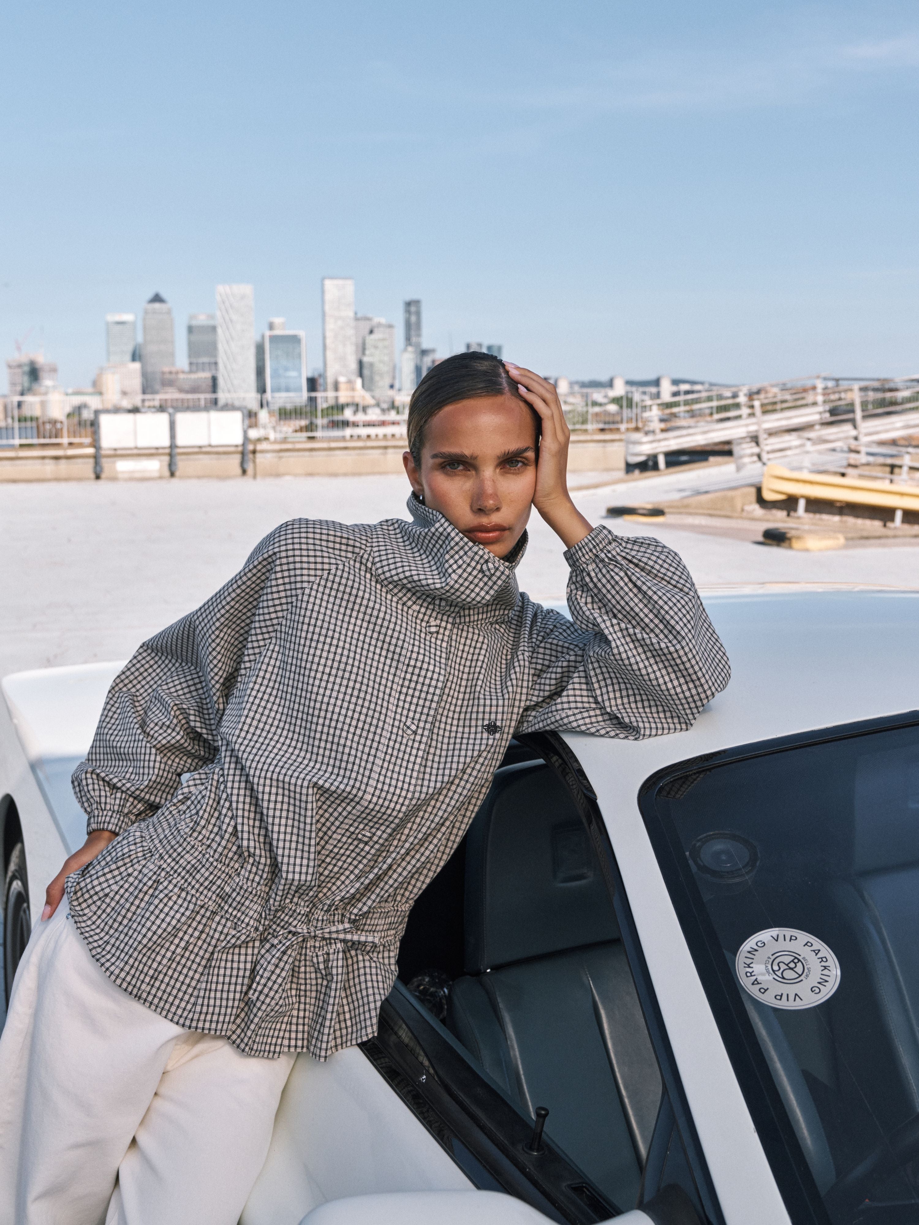 A model wearing a black and white gingham patterned high-neck long-sleeve top and white trousers, leaning against a white car with a city skyline in the background.