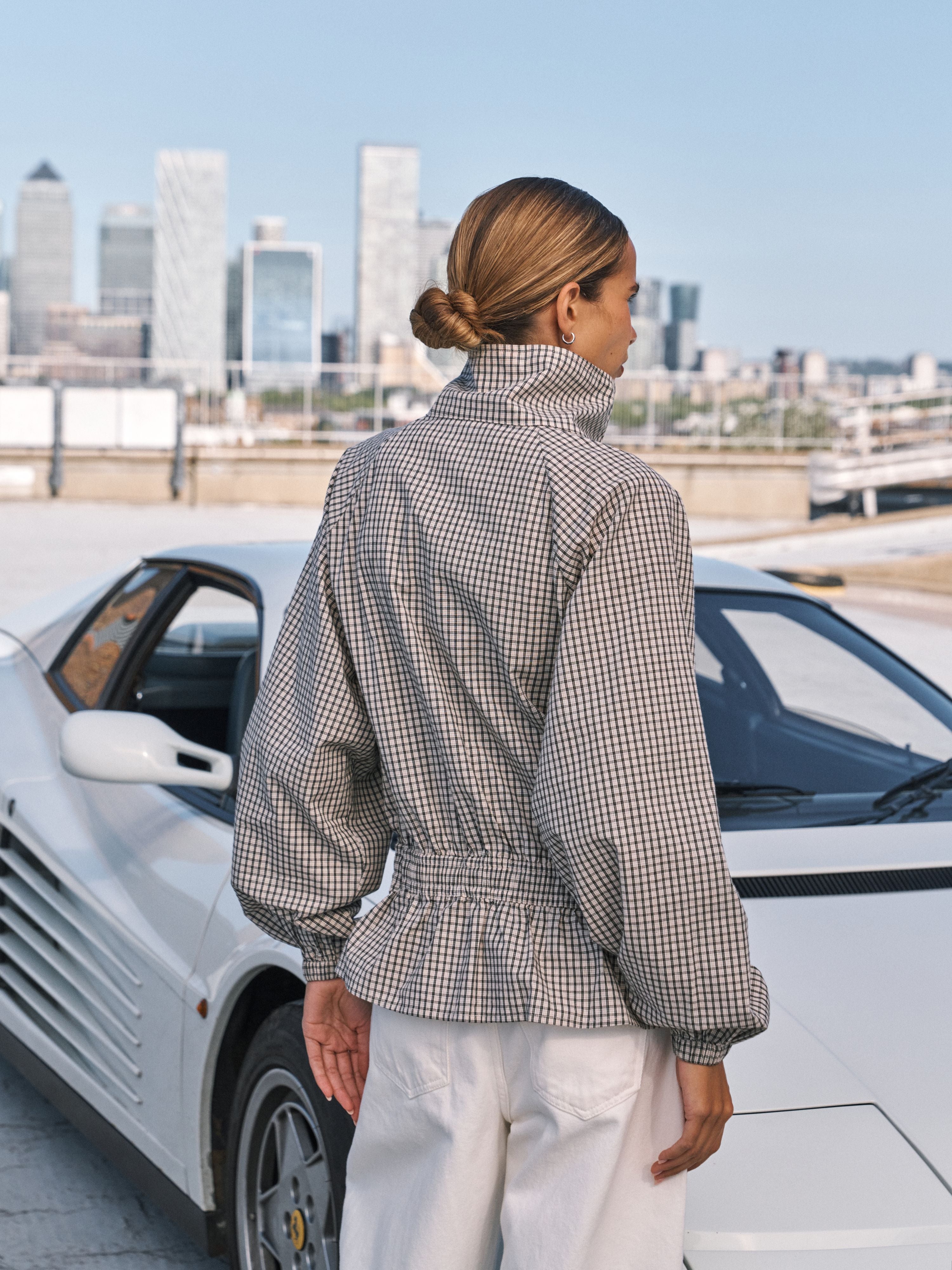 A woman stands outdoors beside a white sports car, shown from the back wearing a high-neck, long-sleeve checkered jacket with a gathered waist and white pants. The background features a city skyline under a clear blue sky.