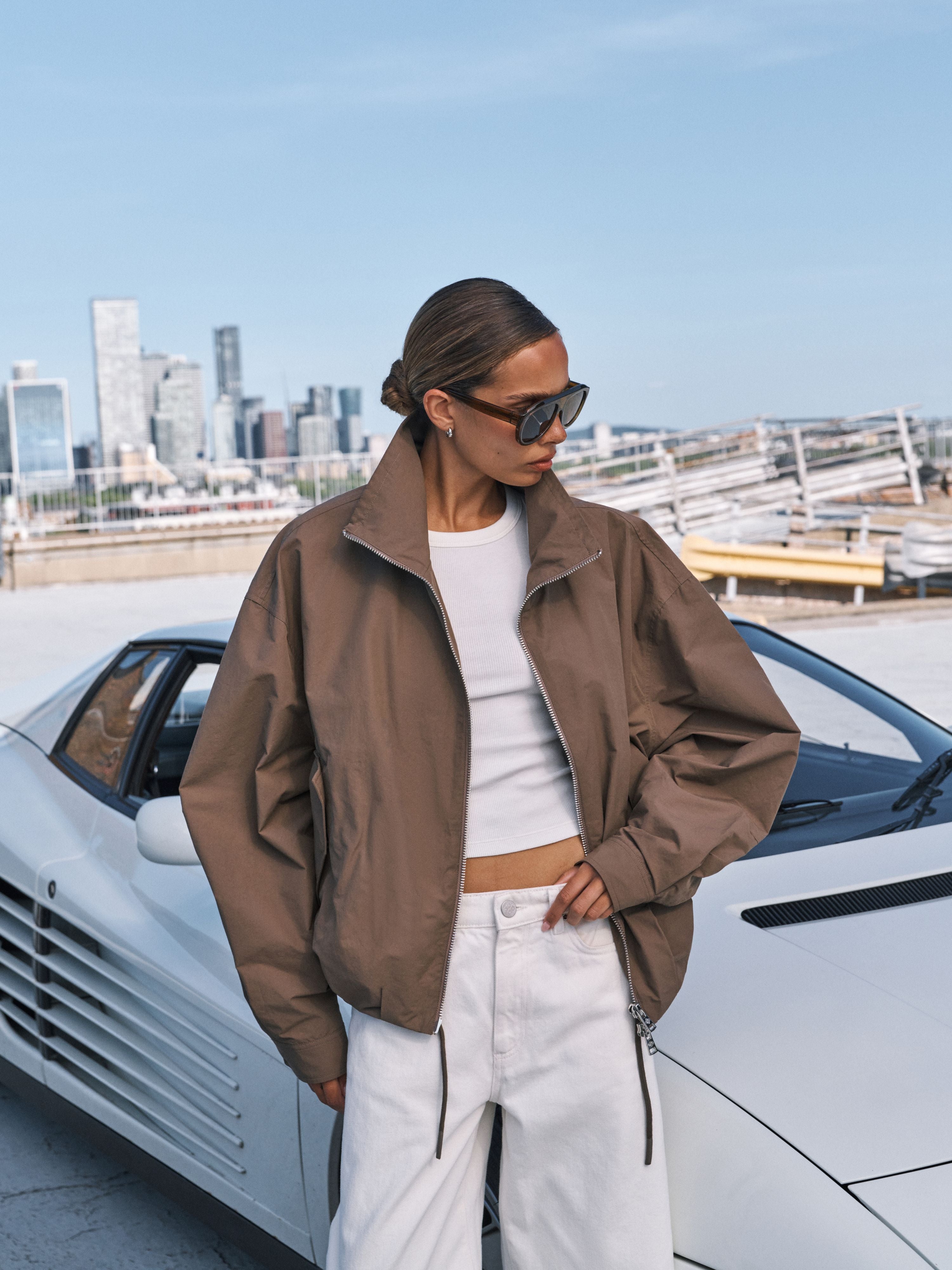 A woman wearing a brown lightweight bomber jacket, white crop top, and white pants stands next to a white sports car with a city skyline in the background.