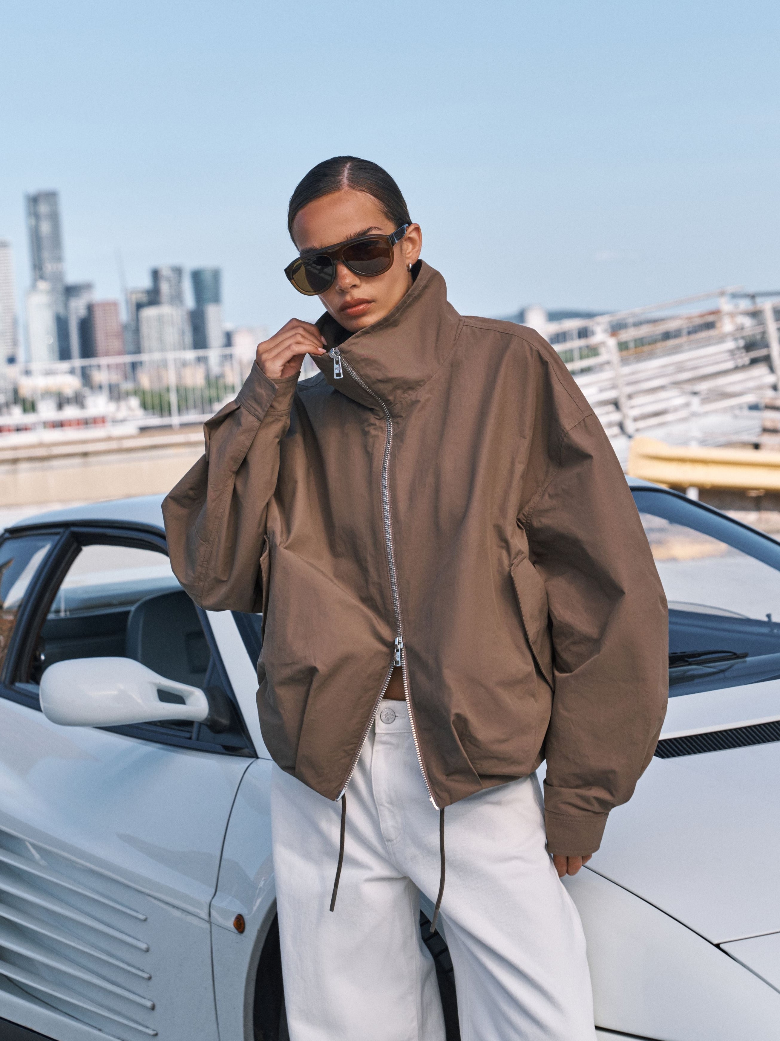 A model wearing a brown zip-up jacket and white pants stands next to a white sports car with a city skyline in the background.