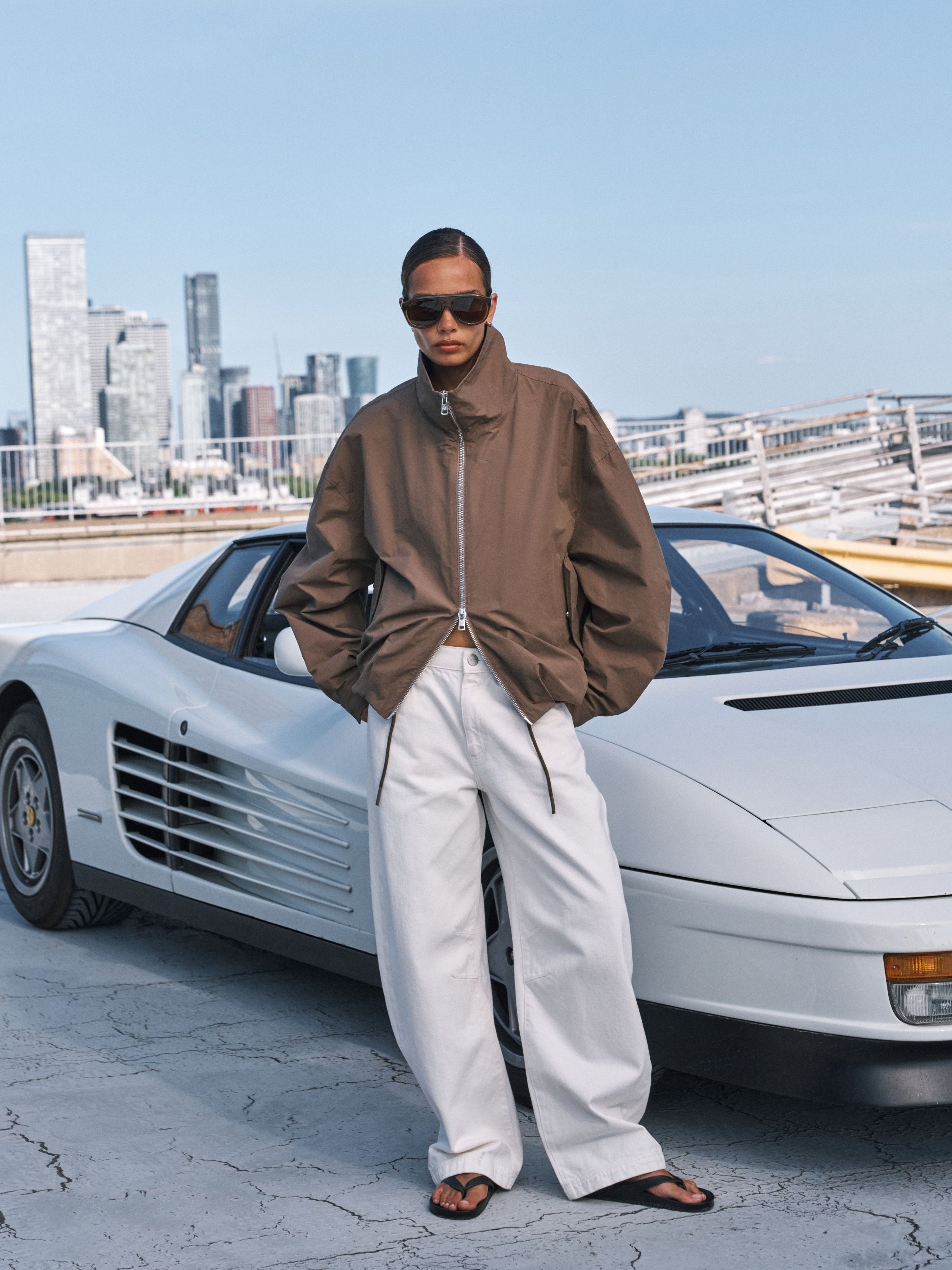 A model stands in front of a white Ferrari Testarossa on a rooftop with a city skyline in the background. The model is wearing a brown zip-up bomber jacket, off-white wide-leg trousers, and dark sunglasses.