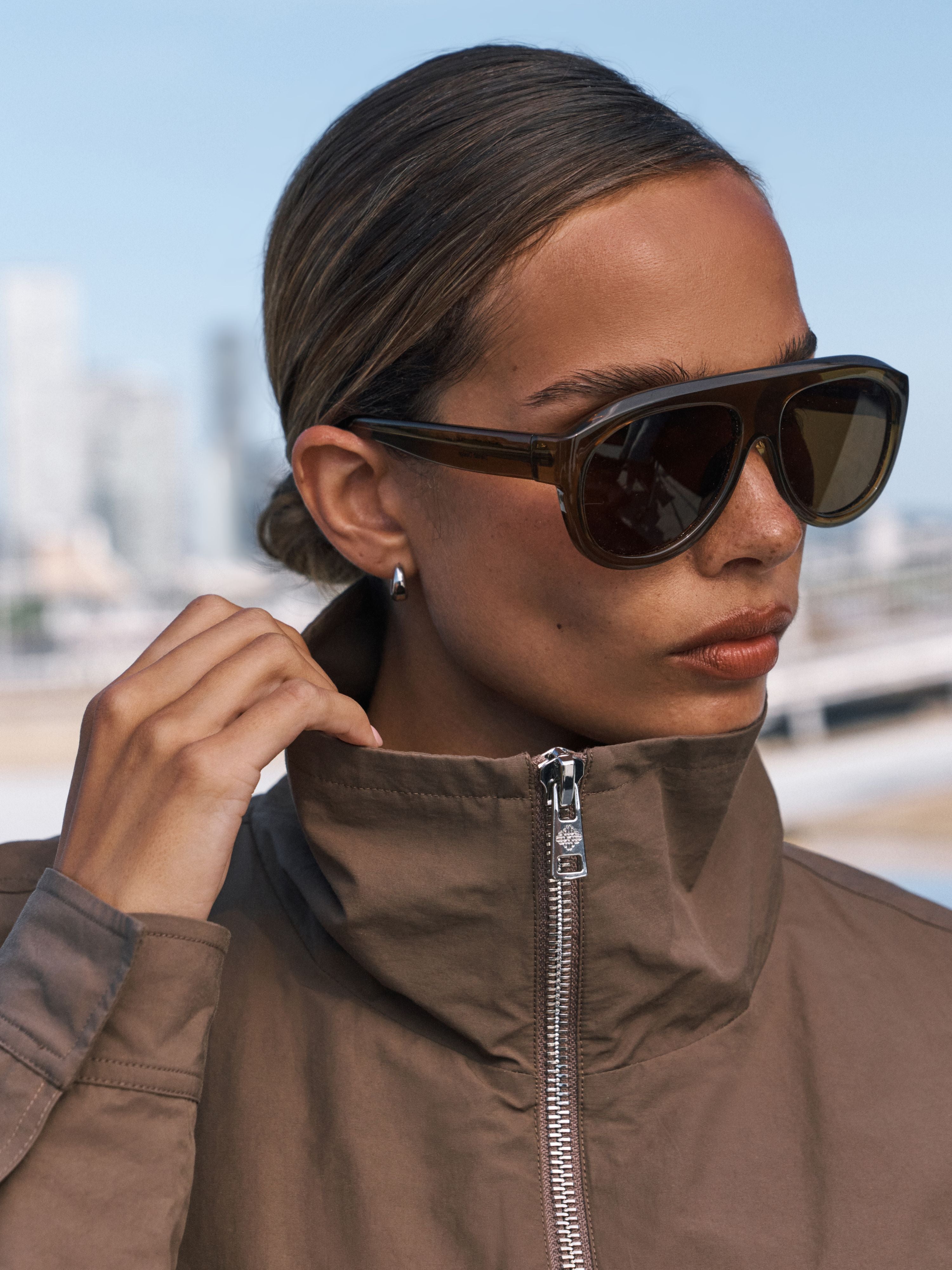 A close-up of a woman wearing dark brown aviator-style sunglasses with a high collar and a brown jacket, set against a blurred outdoor background.